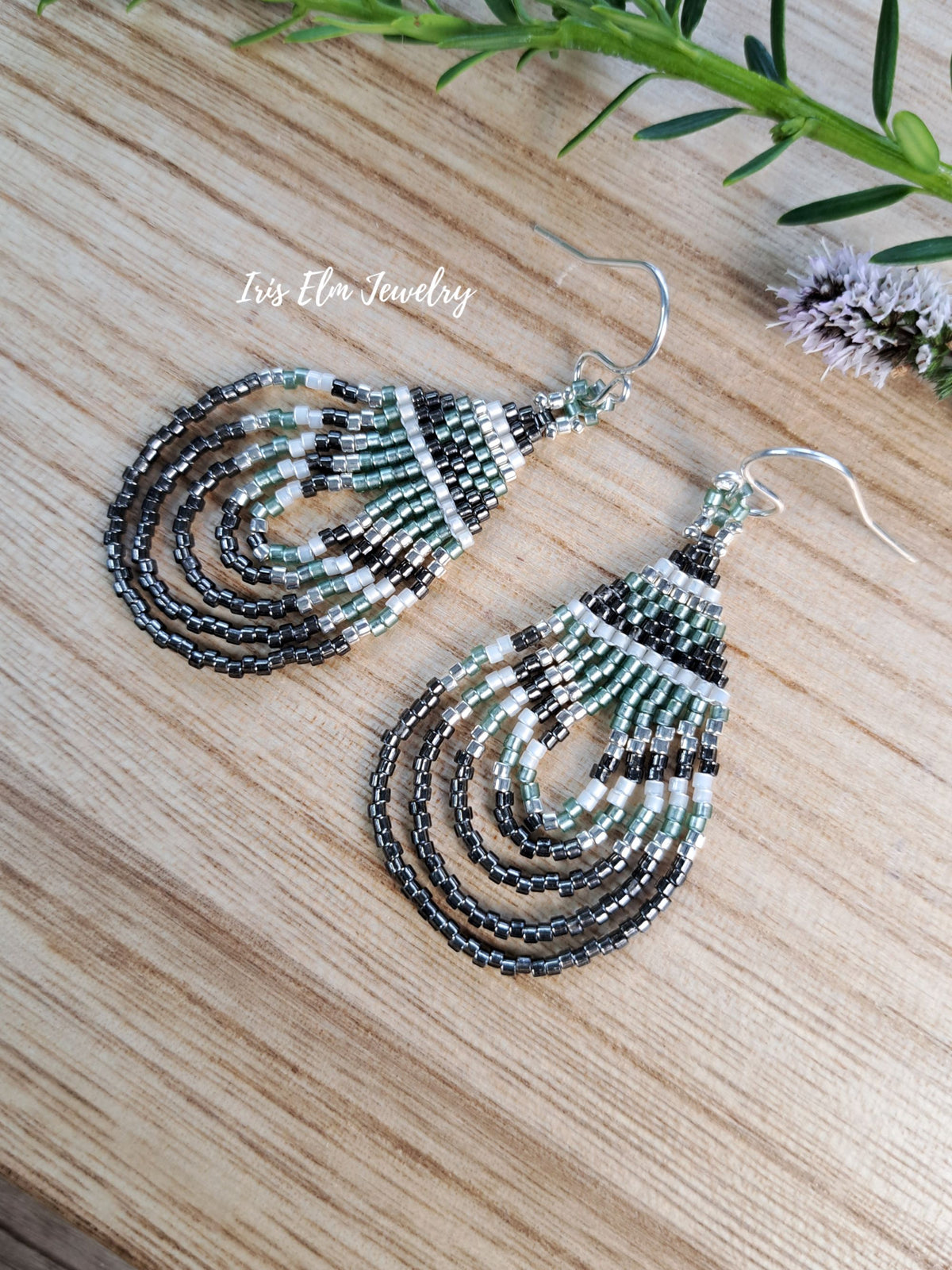 A pair of beaded dangle earrings with silver hooks, featuring concentric teardrop-shaped strands of black, white, and light green seed beads, displayed on a light wood surface with a green sprig in the corner.