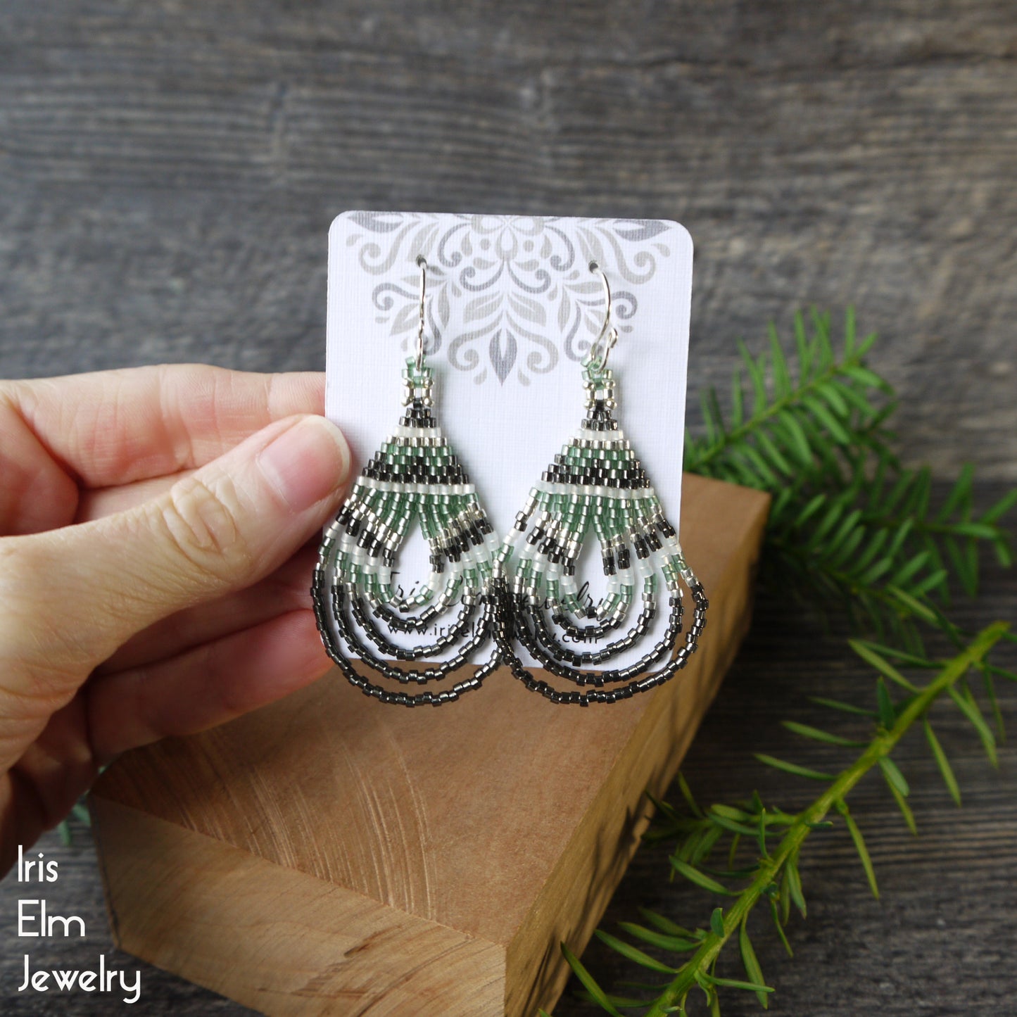 Decorative seed bead glass earrings held by a hand on a wooden block with greenery