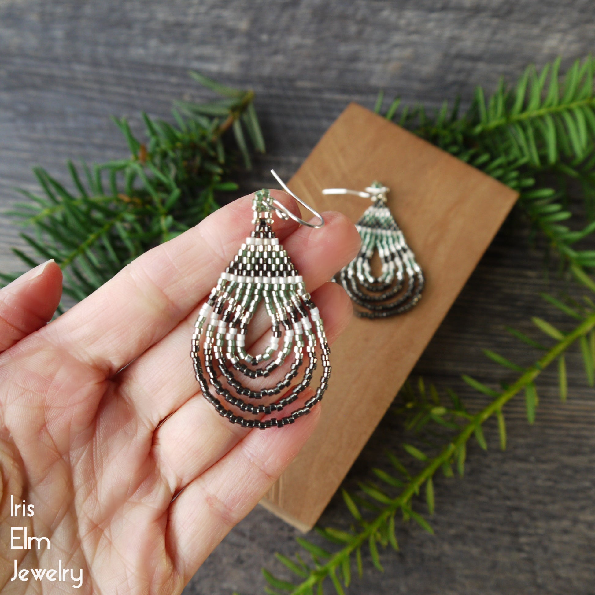 Silver, green, and grey teardrop earrings held in a hand with a wooden block and greenery in the background, featuring 'Iris Elm Jewelry' branding.