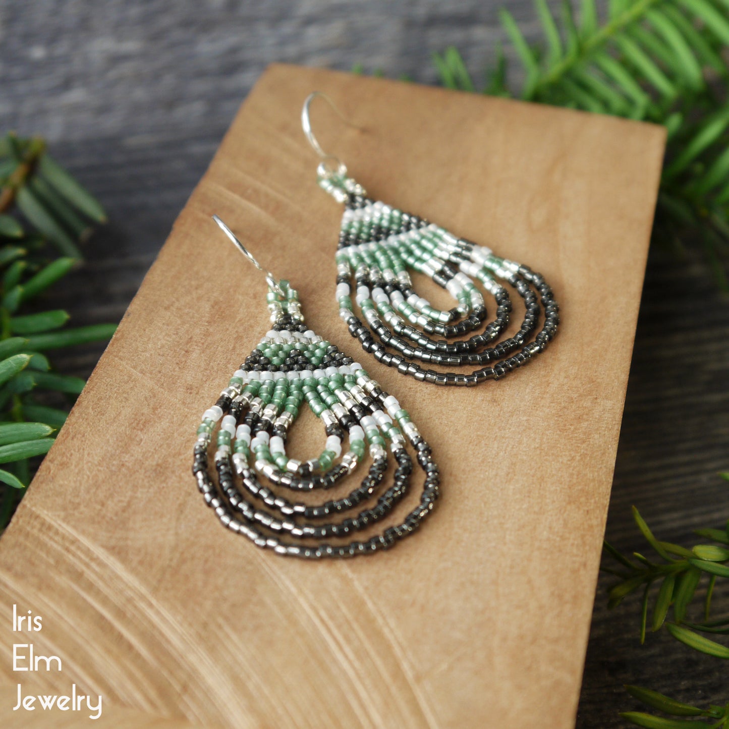 Pair of beaded woven teardrop earrings on a wooden block with greenery in the background, featuring the brand 'Iris Elm Jewelry'.