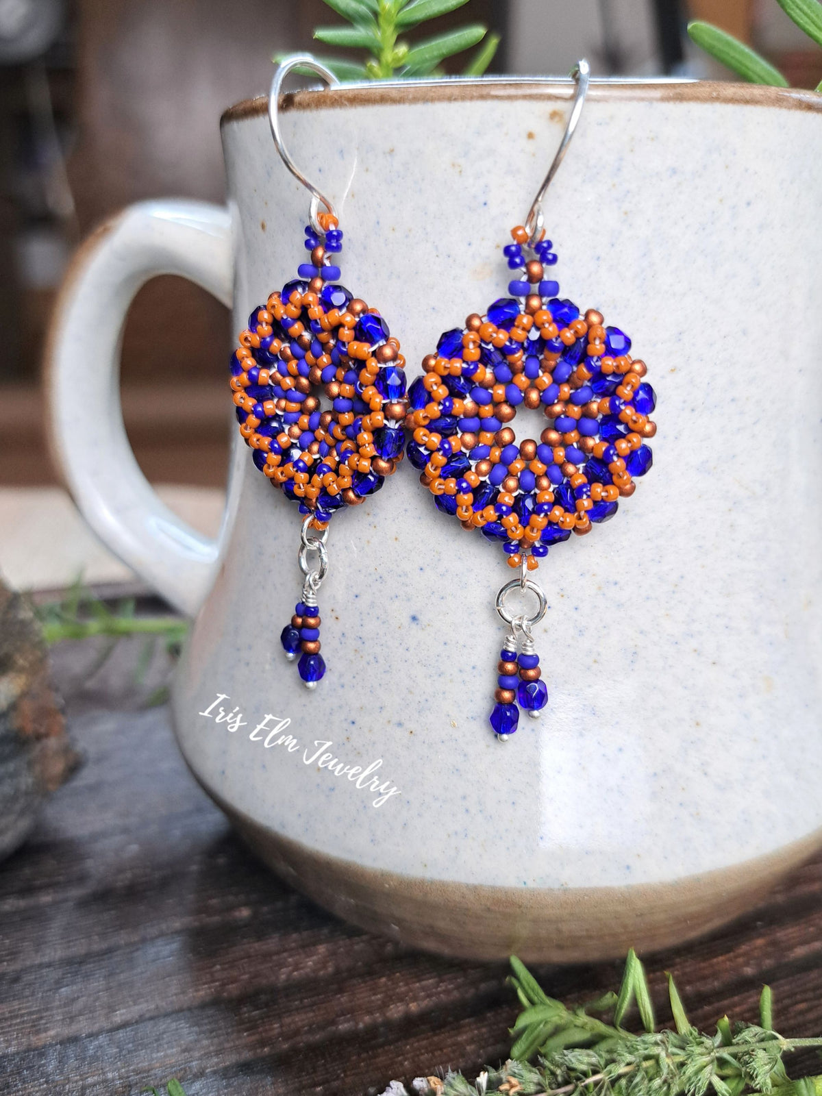 cobalt blue and burnt orange handmade beadwork Czech glass hoop wreath beaded earrings by Iris Elm Jewelry dangling from ceramic mug