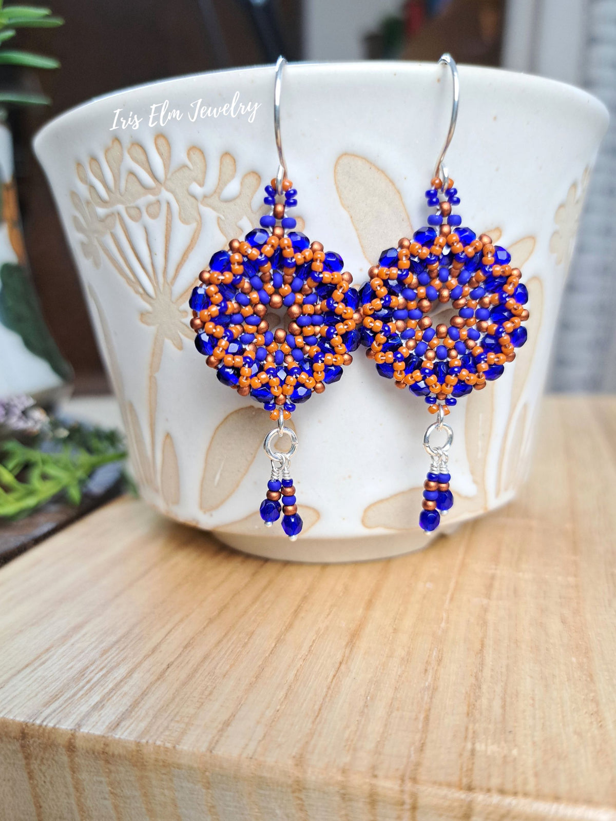 cobalt blue and burnt orange handmade beadwork Czech glass hoop wreath beaded earrings by Iris Elm Jewelry dangling from ceramic mug
