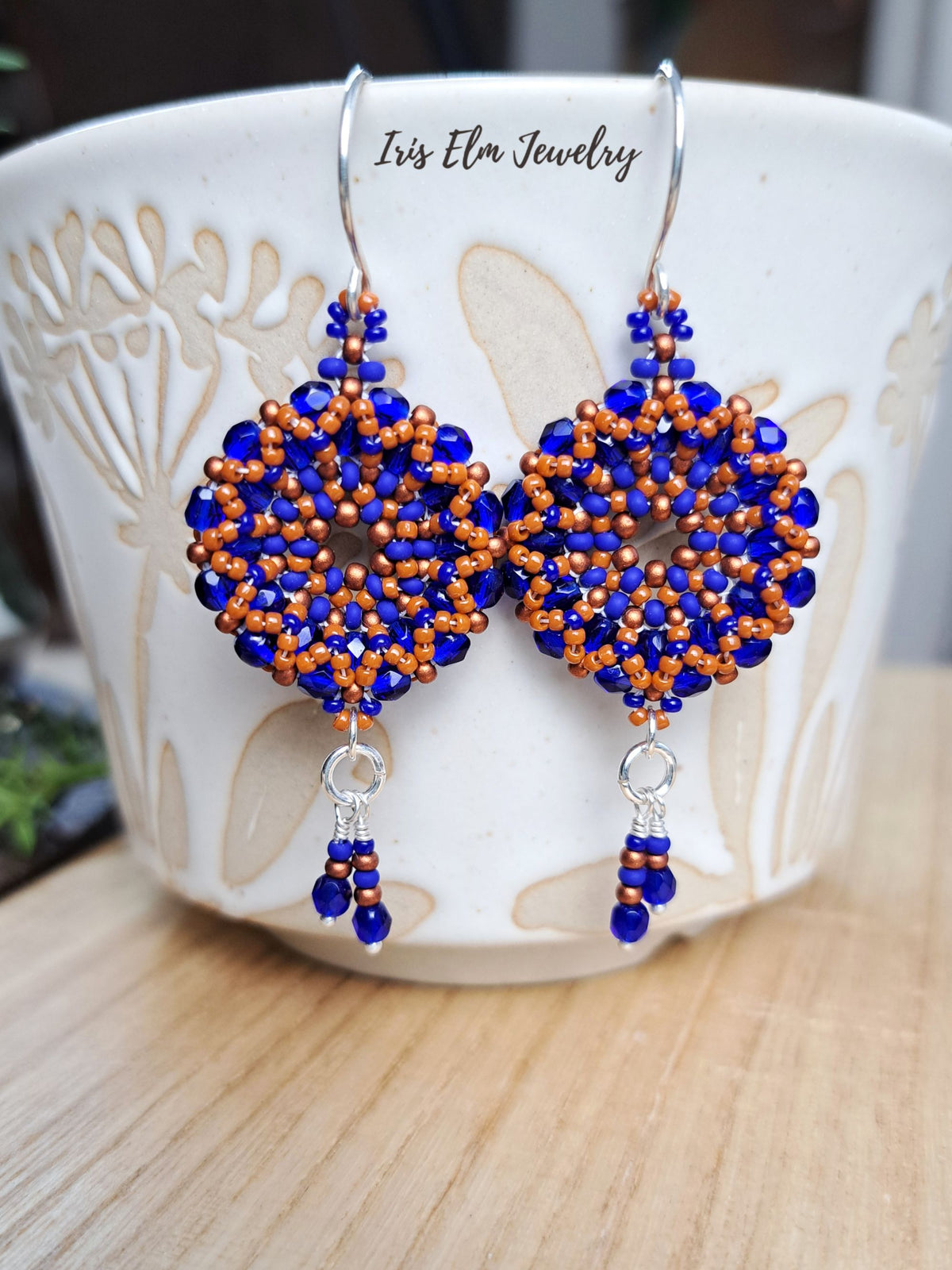cobalt blue and burnt orange handmade beadwork Czech glass hoop wreath beaded earrings by Iris Elm Jewelry dangling from ceramic mug