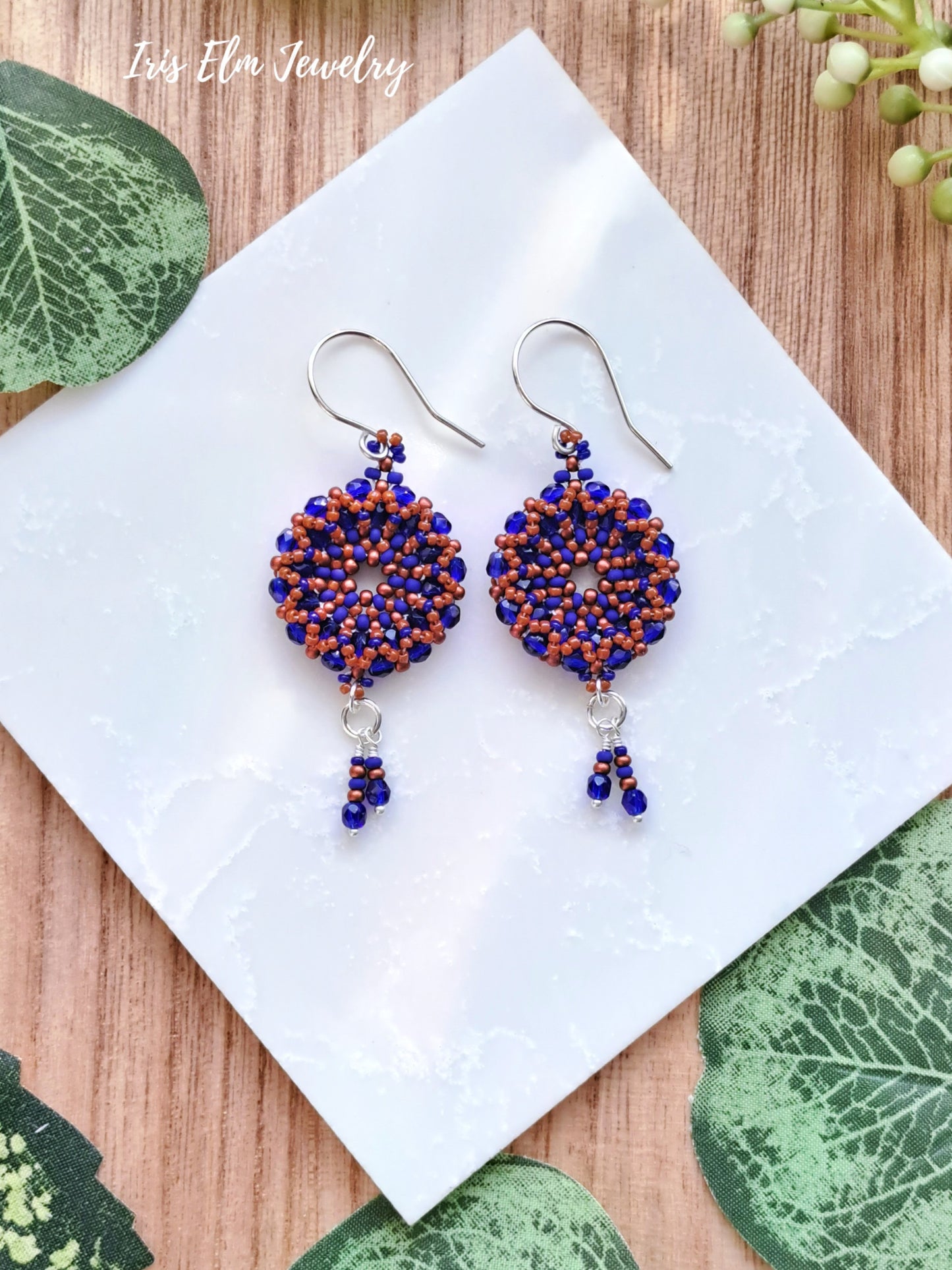 Pair of beaded cobalt blue and burnt orange woven glass wreath hoop earrings on a white square with green leaves around
