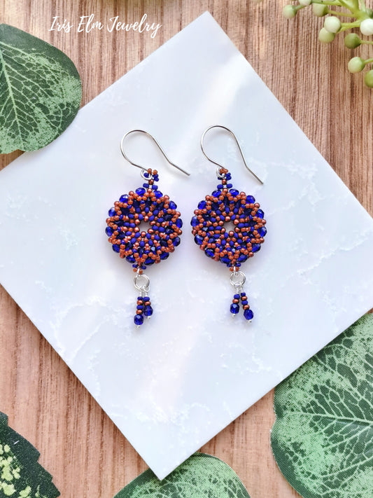 Pair of beaded cobalt blue and burnt orange woven glass wreath hoop earrings on a white square with green leaves around
