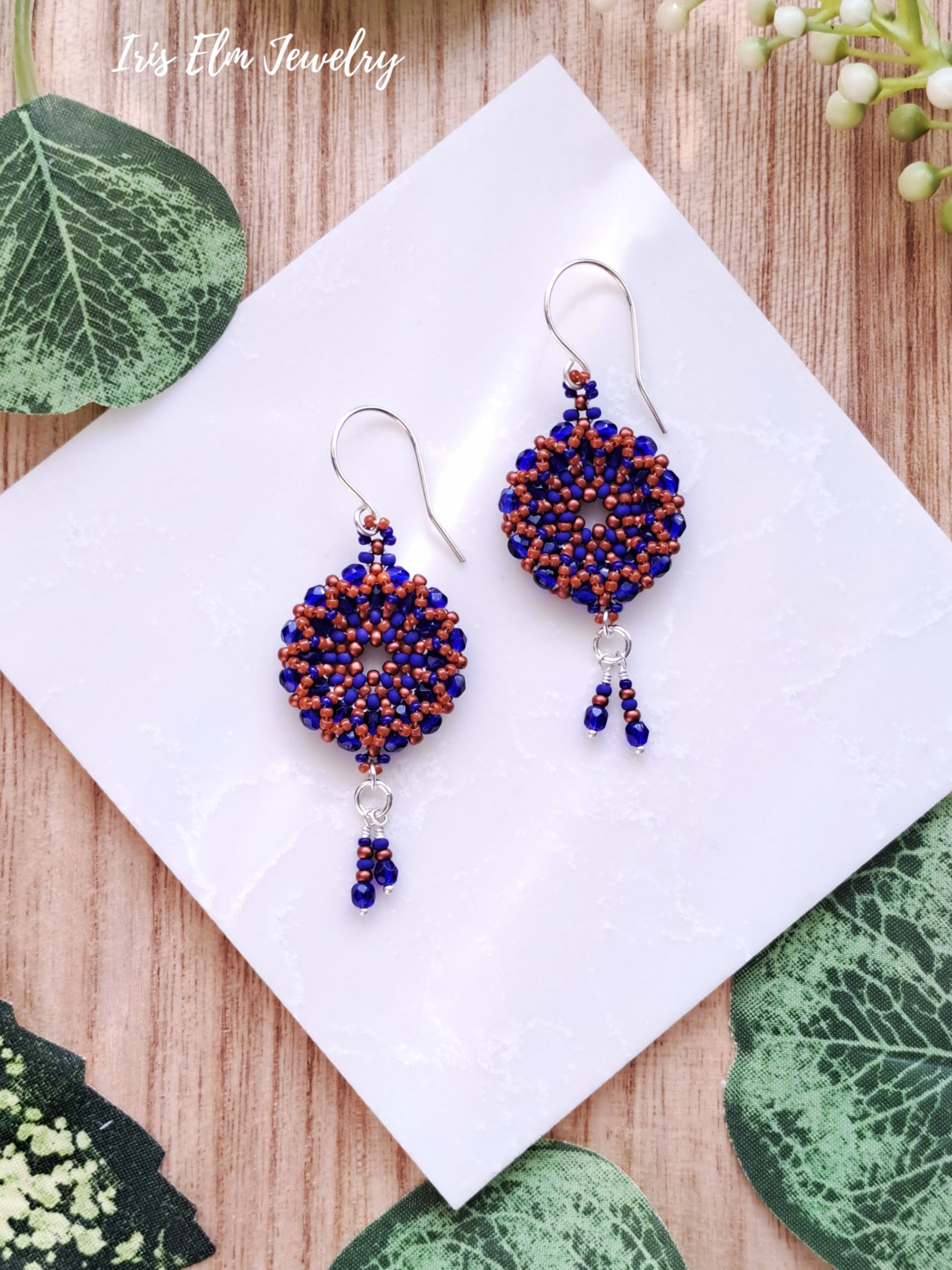 Pair of beaded cobalt blue and burnt orange woven glass wreath hoop earrings on a white tile with green leaves in the background