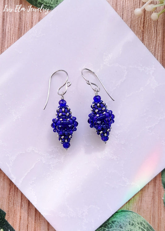 Pair of cobalt blue glass beaded bead earrings on a white napkin with 'Iris Elm Jewelry' branding.