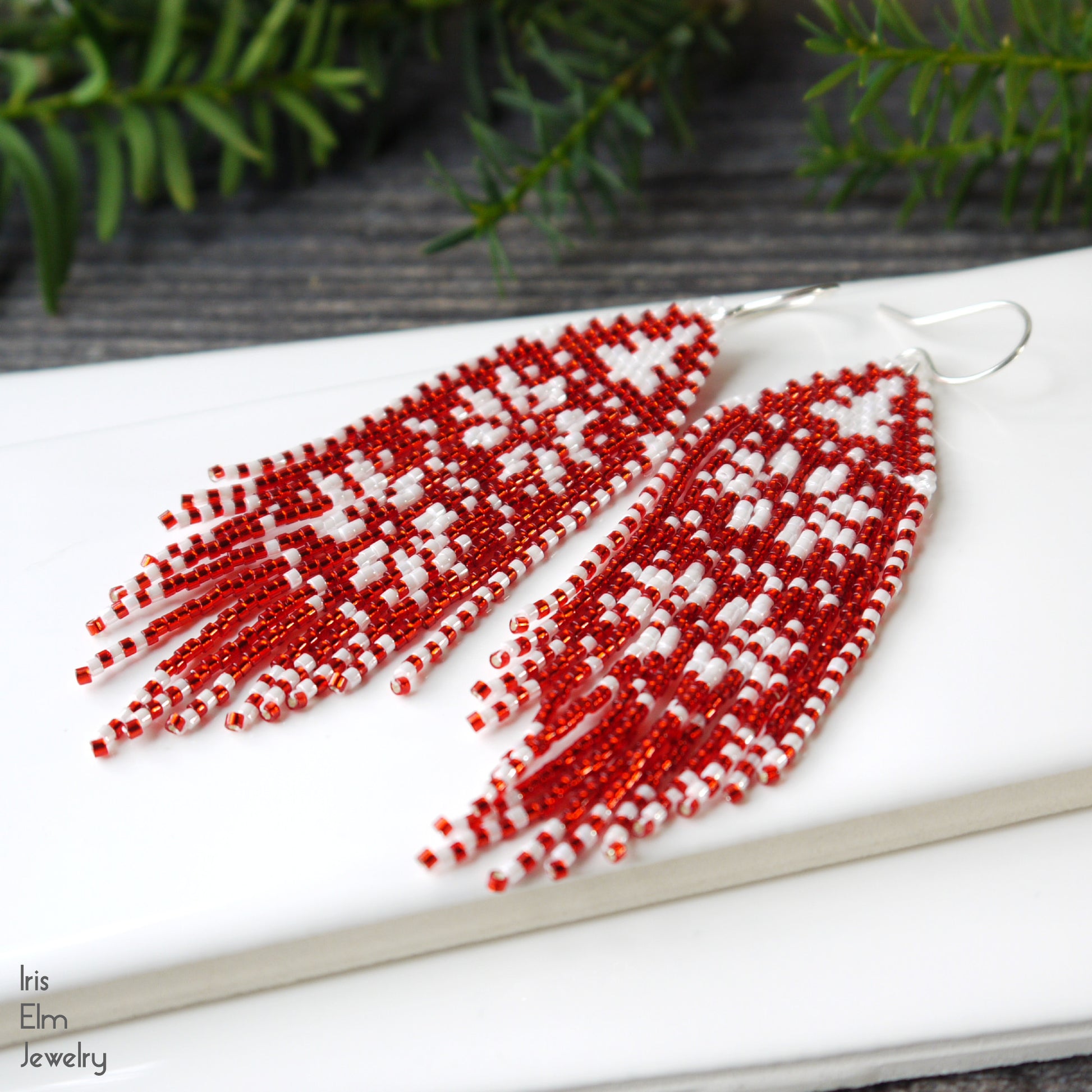 Red and white beaded winter tree and snowflake fringe seed bead earrings on a white surface with greenery in the background