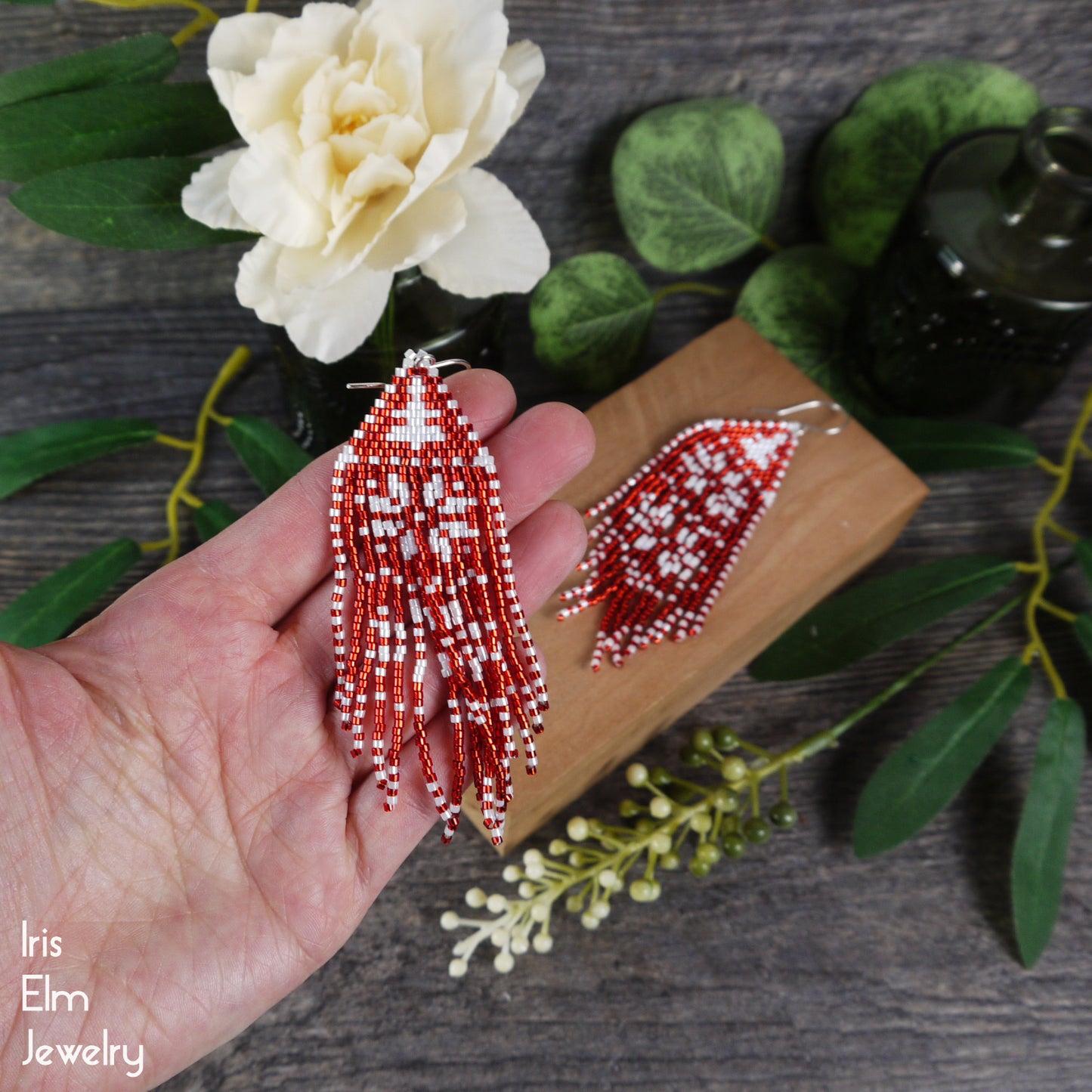 Beaded red and white snowflake seed bead fringe earrings held in a hand with a wooden block and flowers in the background, featuring 'Iris Elm Jewelry'.