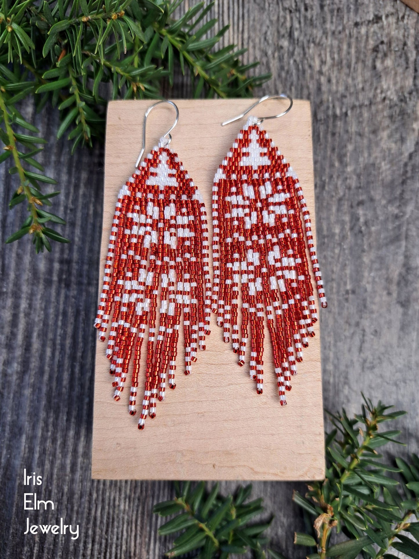 Red and white beaded fringe snowflake earrings on a wooden display with greenery in the background