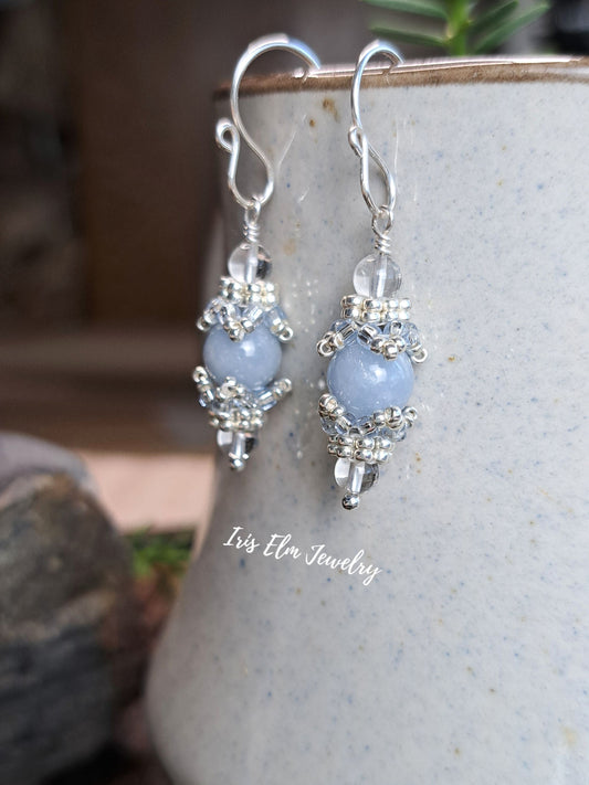 A pair of handmade silver dangle earrings with light blue angelite beads and clear crystal accents, shown hanging from the rim of a speckled ceramic mug. The brand name 'Iris Elm Jewelry' is watermarked on the front.