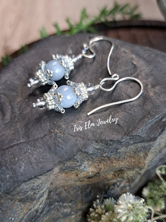 A pair of silver-toned dangle earrings with light blue angelite beads and clear quartz crystal  accents, displayed on a dark textured stone surface with a visible 'Iris Elm Jewelry' watermark.