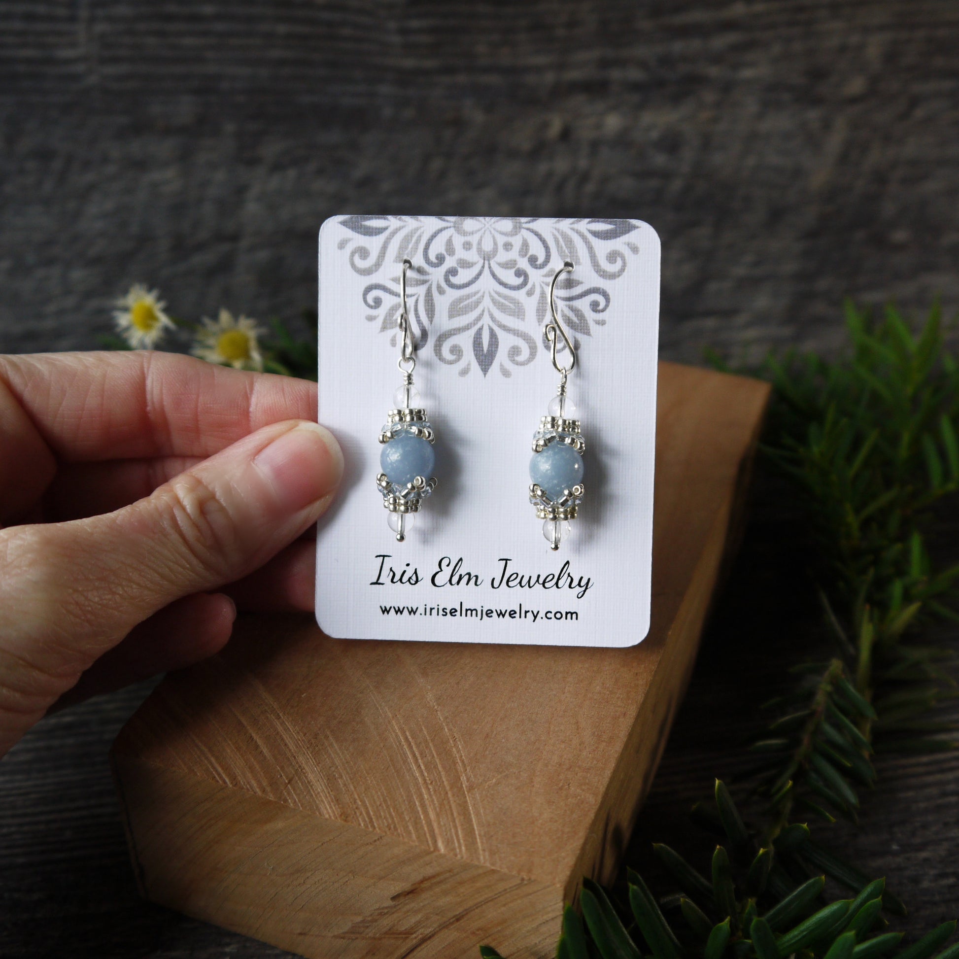 Pair of blue angelite handmade earrings on a 'Iris Elm Jewelry' card held by a hand with a natural background.