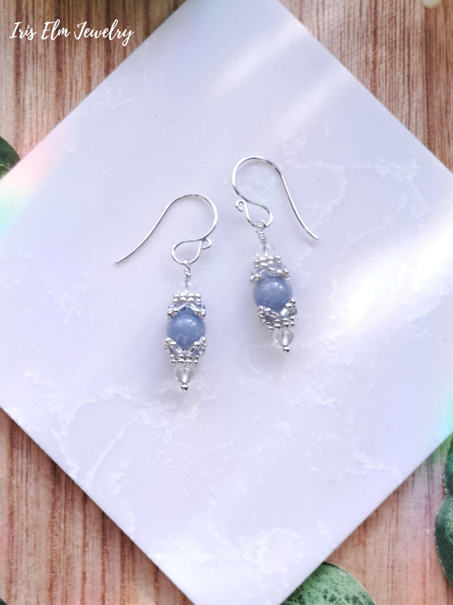 Pair of silver earrings with blue angelite beads on a white cloth with 'Iris Elm Jewelry' branding.