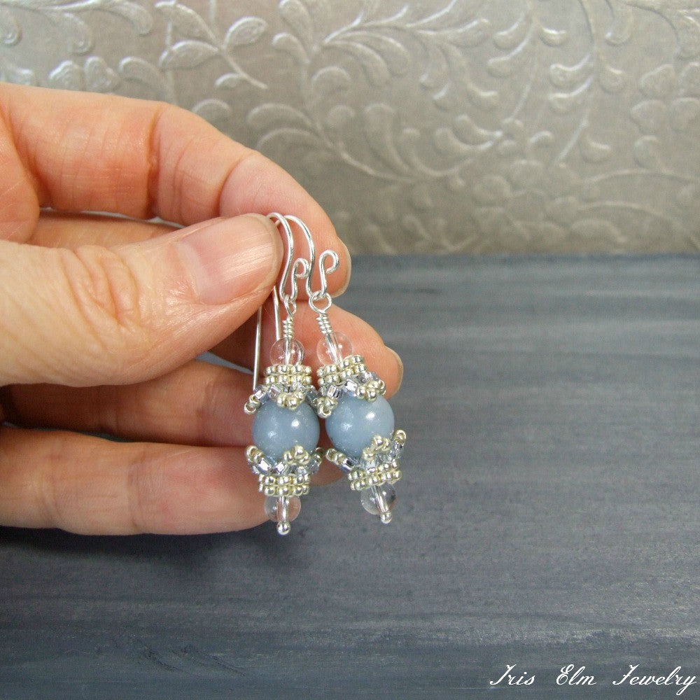 Hand holding a pair of blue angelite and quartz beaded earrings with a textured gray background