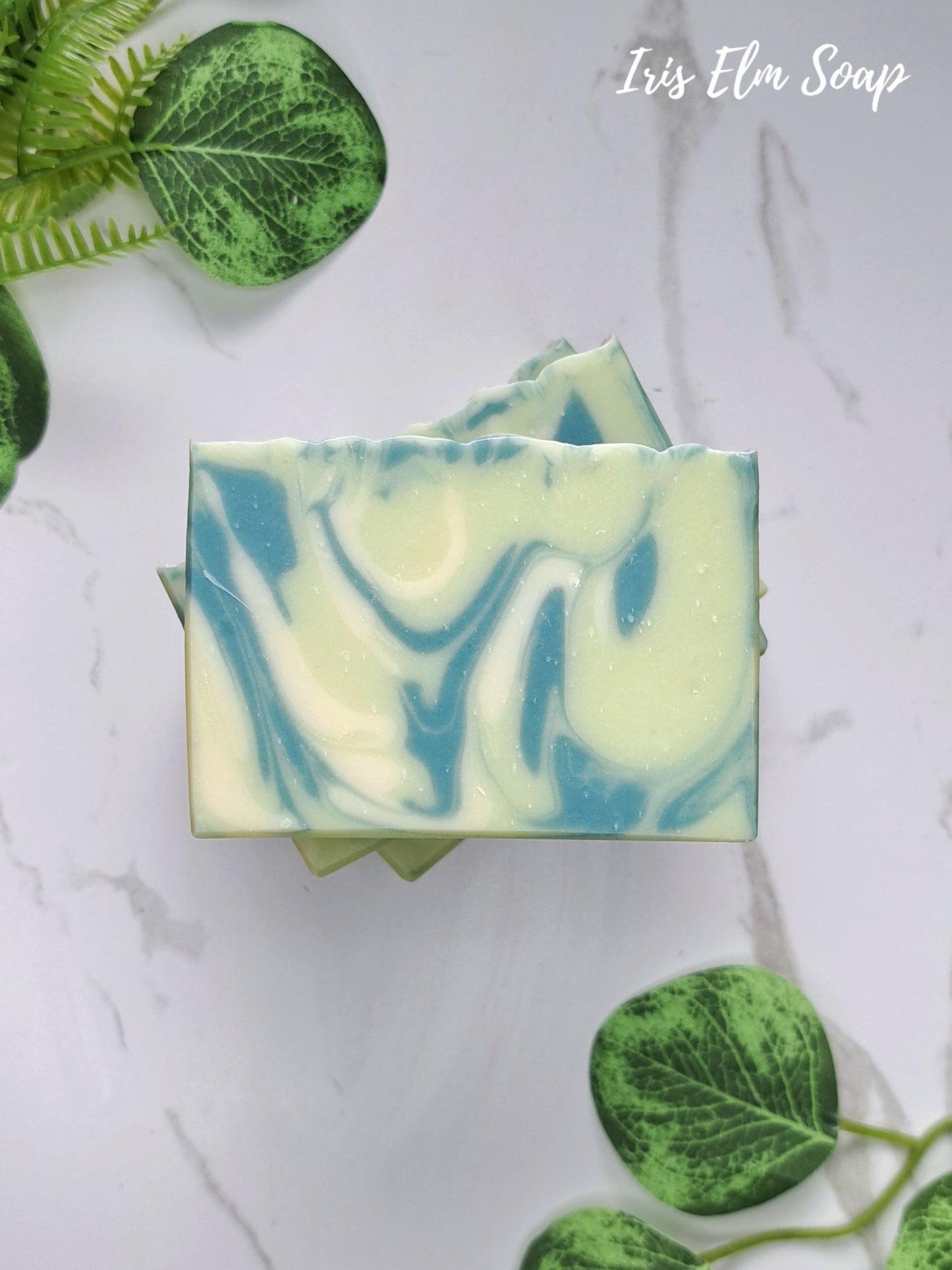 A rectangular bar of marbled agave lime handmade soap with swirls of light blue and pale yellow-green, stacked on a white marble surface with green leafy stems around it. The text 'Iris Elm Soap' appears in the top right.
