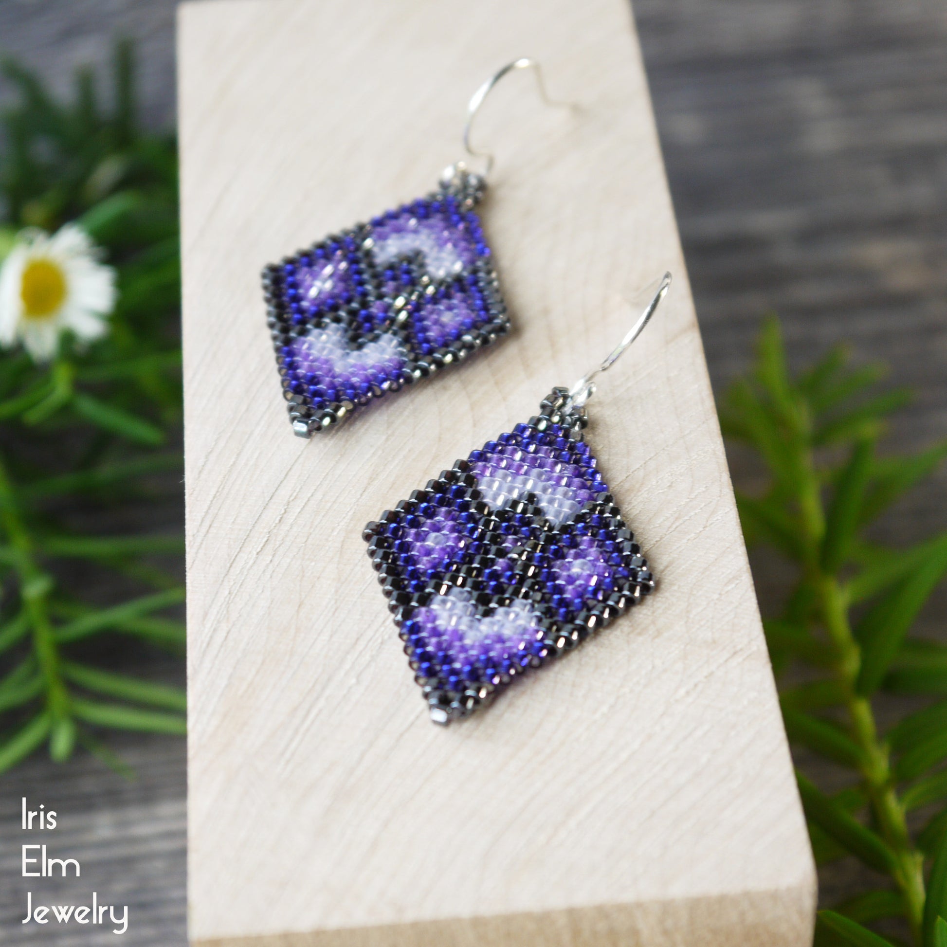 Pair of purple beaded diamond shape seed bead earrings on a wooden block with greenery in the background, featuring 'Iris Elm Jewelry' branding.