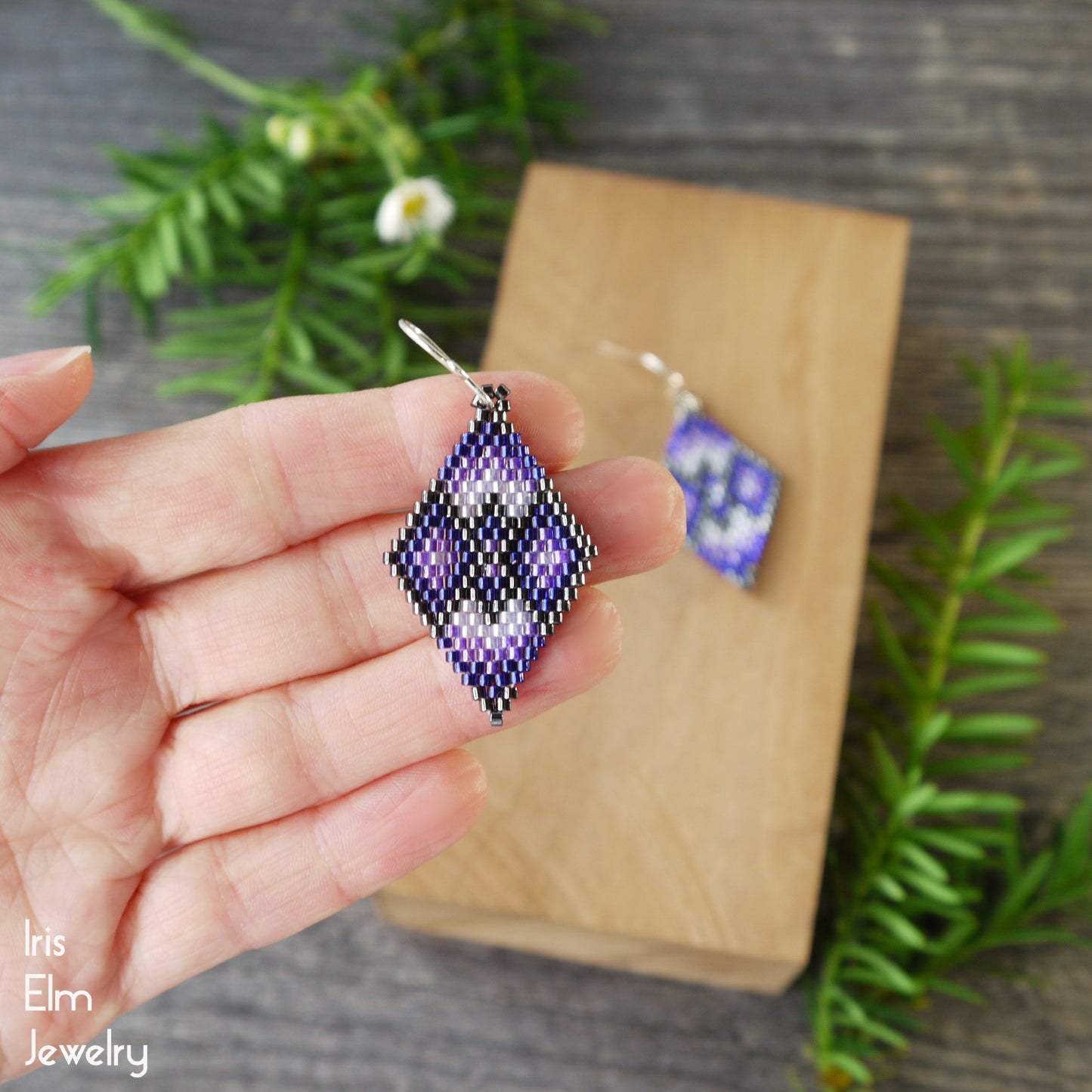 Hand holding a beaded purple woven earring with 'Iris Elm Jewelry' branding, set against a wooden block and greenery.