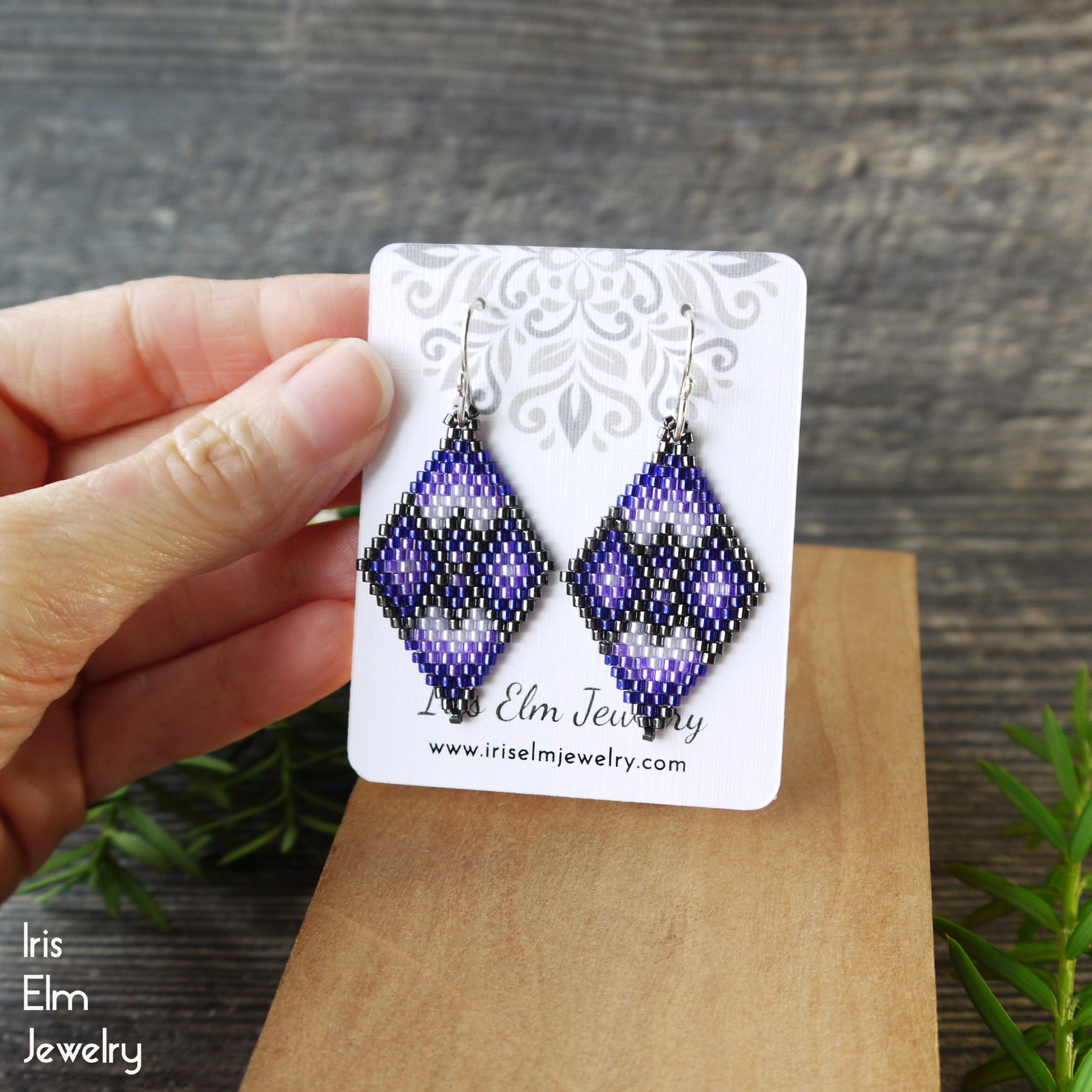 Pair of purple geometric beaded earrings on a 'Iris Elm Jewelry' card held by a hand with a wooden background.