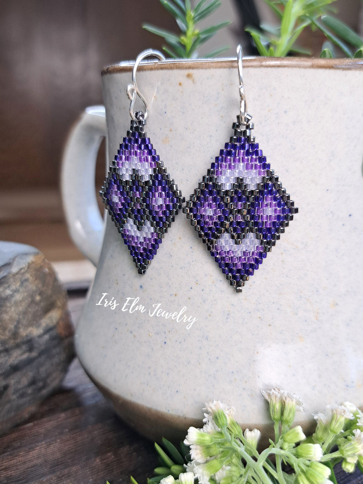 A pair of handmade beaded dangle earrings with a geometric diamond pattern in purple, black, and white, hanging from silver fishhook wires. The earrings are displayed in front of a speckled beige ceramic mug with green sprigs and small white flowers.