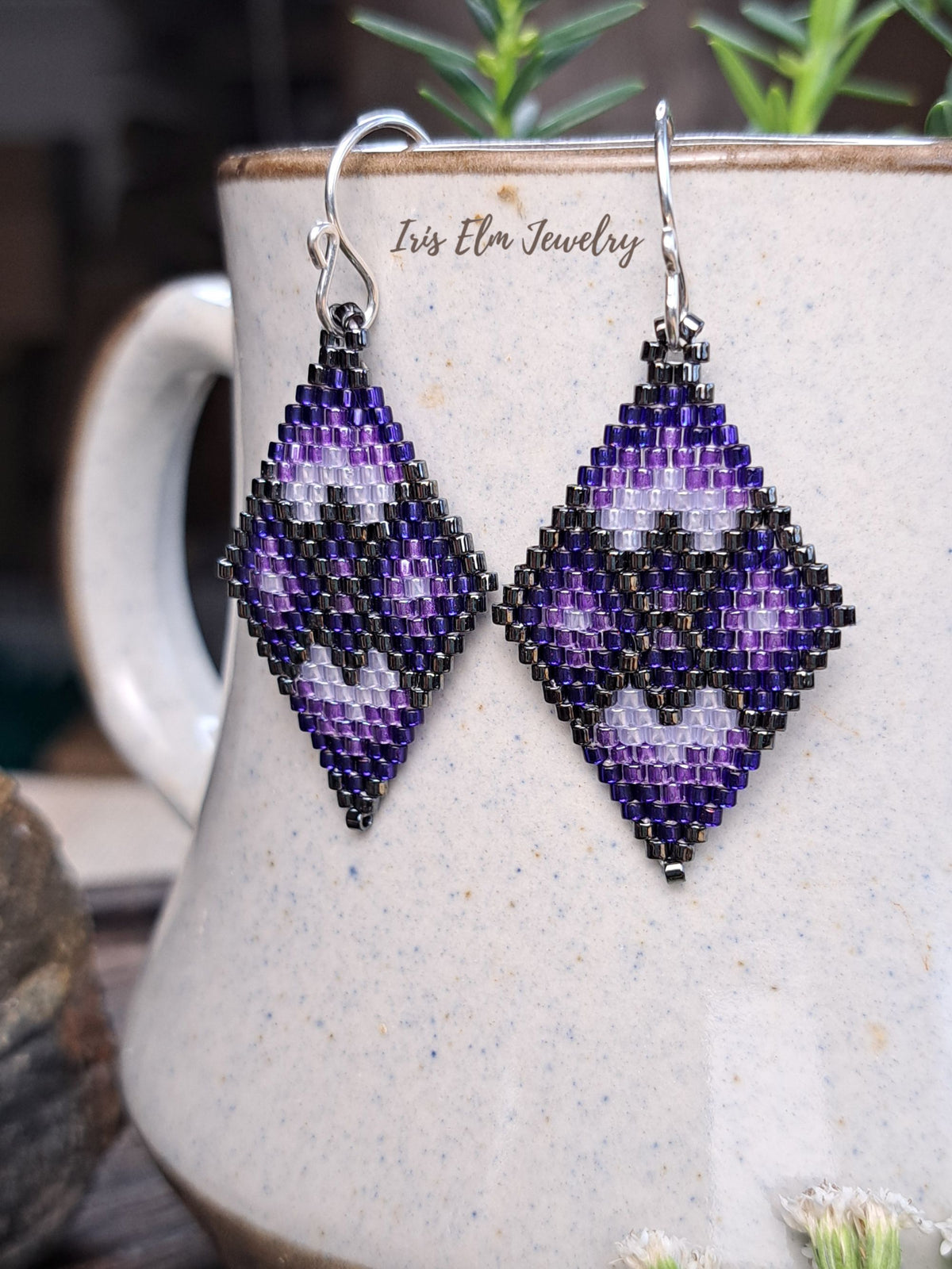 A pair of handmade beaded dangle earrings with a geometric diamond pattern in purple, black, and white, hanging from silver hooks. They are displayed against a speckled ceramic mug with the brand name 'Iris Elm Jewelry' printed on it, with blurred green foliage in the background.