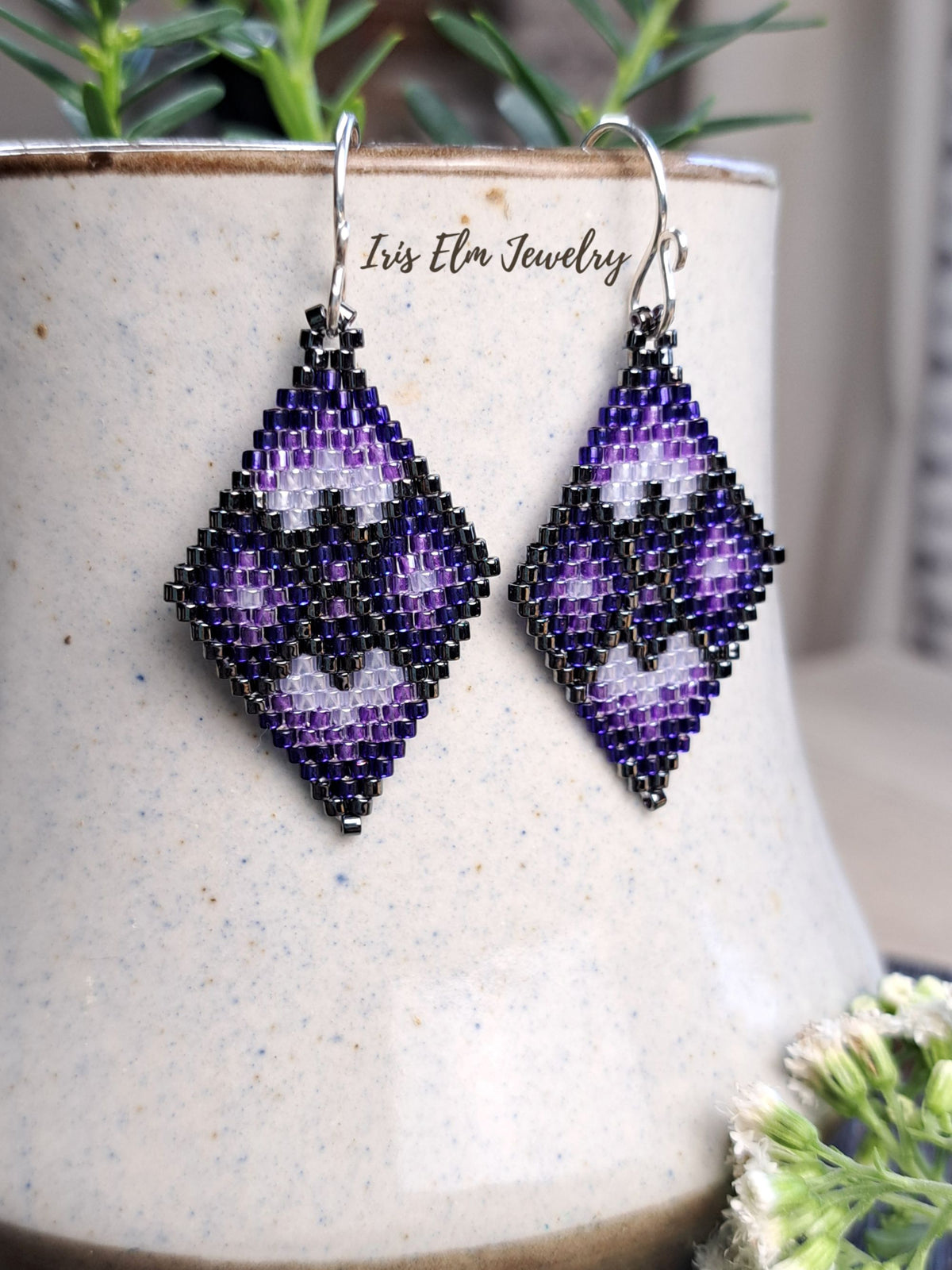 A pair of handmade beaded dangle earrings with a purple and grey geometric pattern, hanging from silver fishhook wires. The earrings are displayed against a speckled beige ceramic pot with green foliage in the background.
