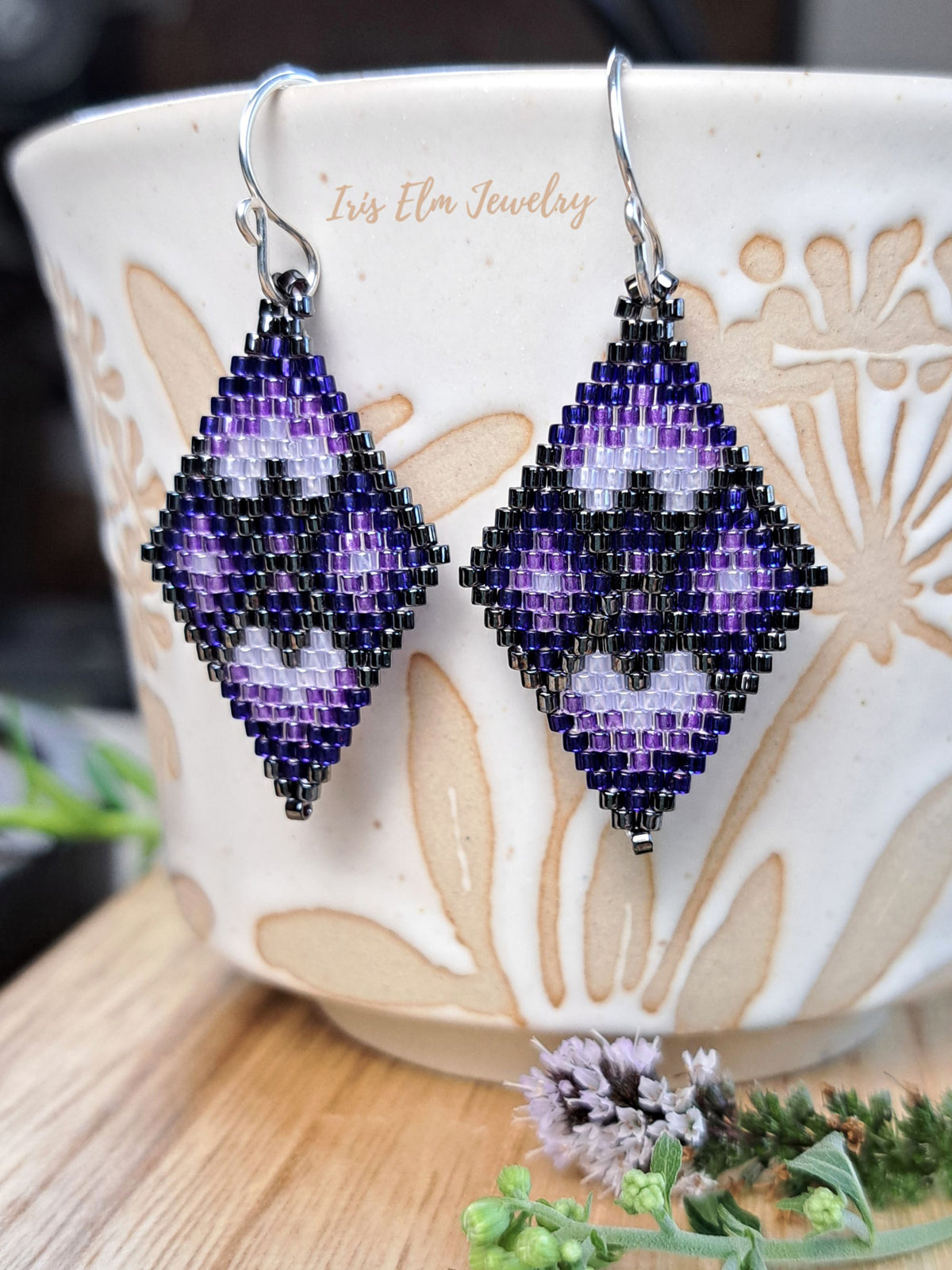 A pair of handmade beaded dangle earrings with a purple, black, and white geometric pattern, hanging in front of a ceramic cup. The brand name 'Iris Elm Jewelry' is printed on the cup.