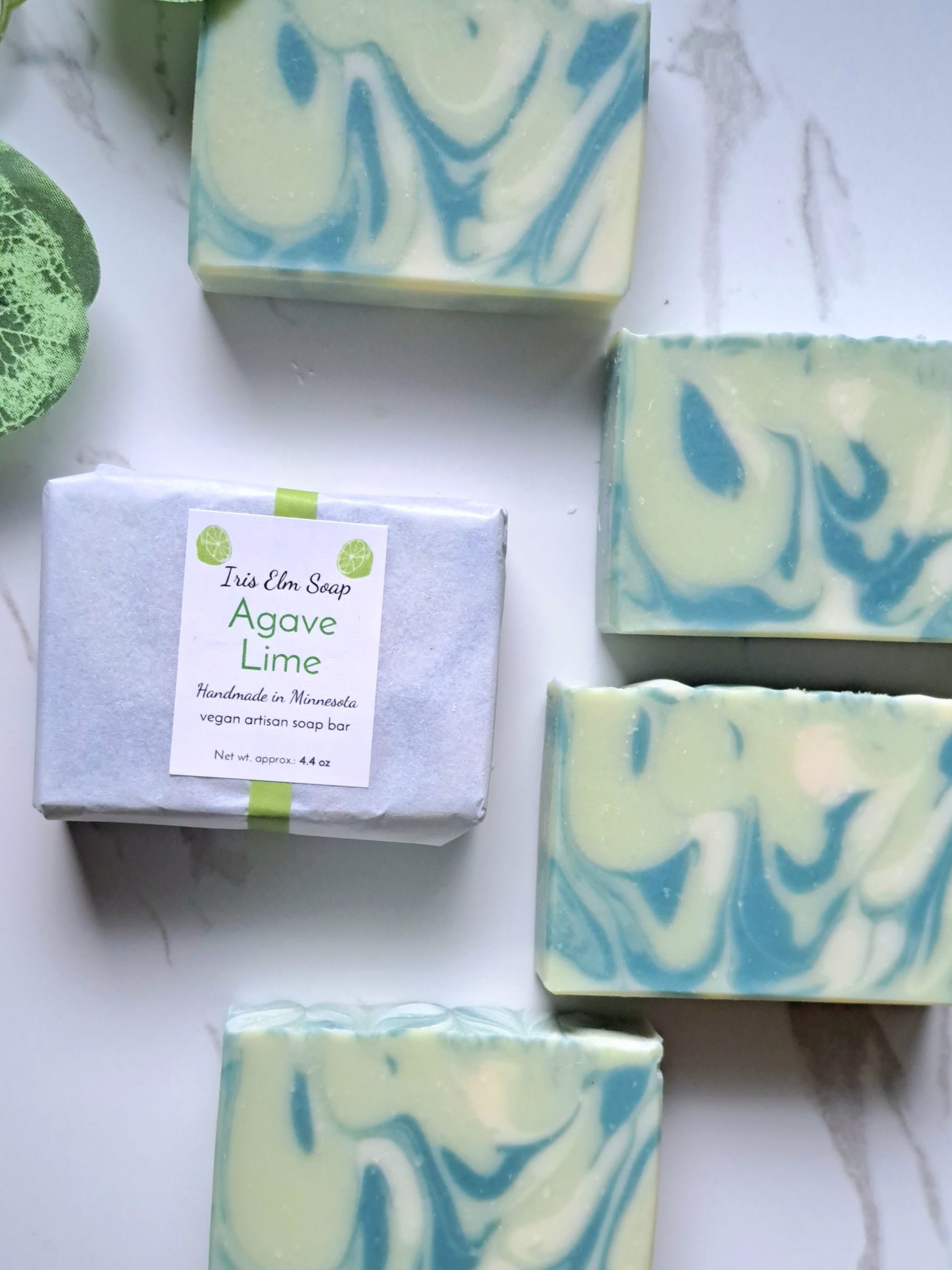 Five bars of artisan soap displayed on a white marble surface, including one wrapped bar labeled 'Iris Elm Soap Agave Lime'. The soaps feature light green, green, and white marbled swirls.