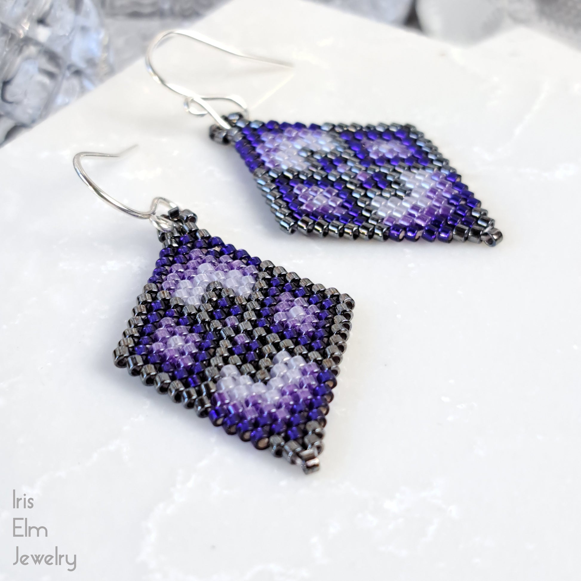 Pair of beaded purple diamond shape seed bead earrings with a geometric pattern on a white surface, branded 'Iris Elm Jewelry'.