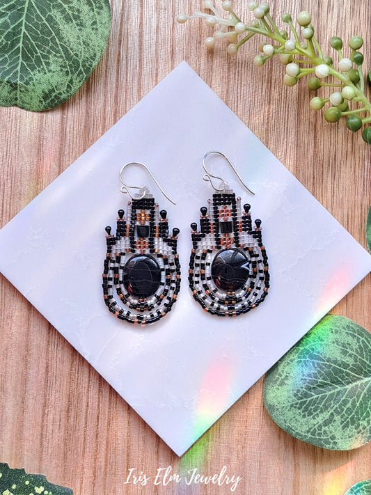 Pair of black and white agate stone and seed bead beaded earrings on a white card with green leaves in the background