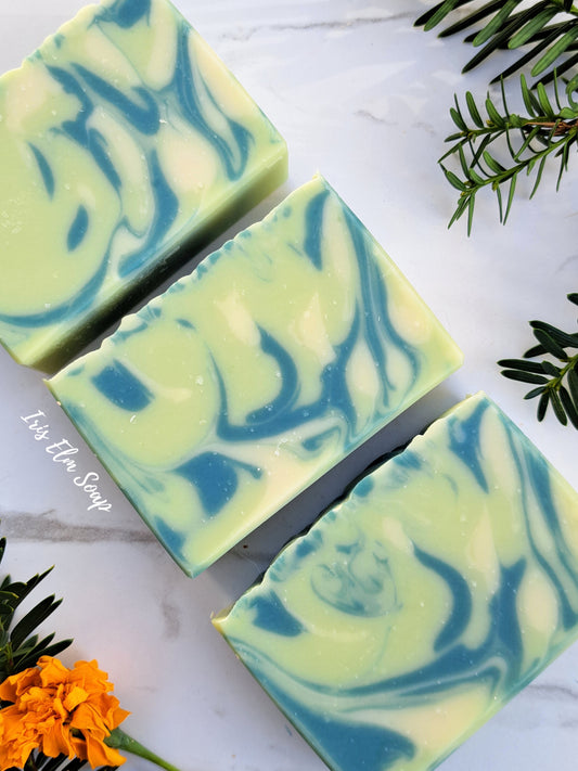 Three rectangular bars of agave lime handmade soap with a blue and pale green marbled swirl pattern, arranged diagonally on a white marble surface with decorative green leaves and orange flowers.