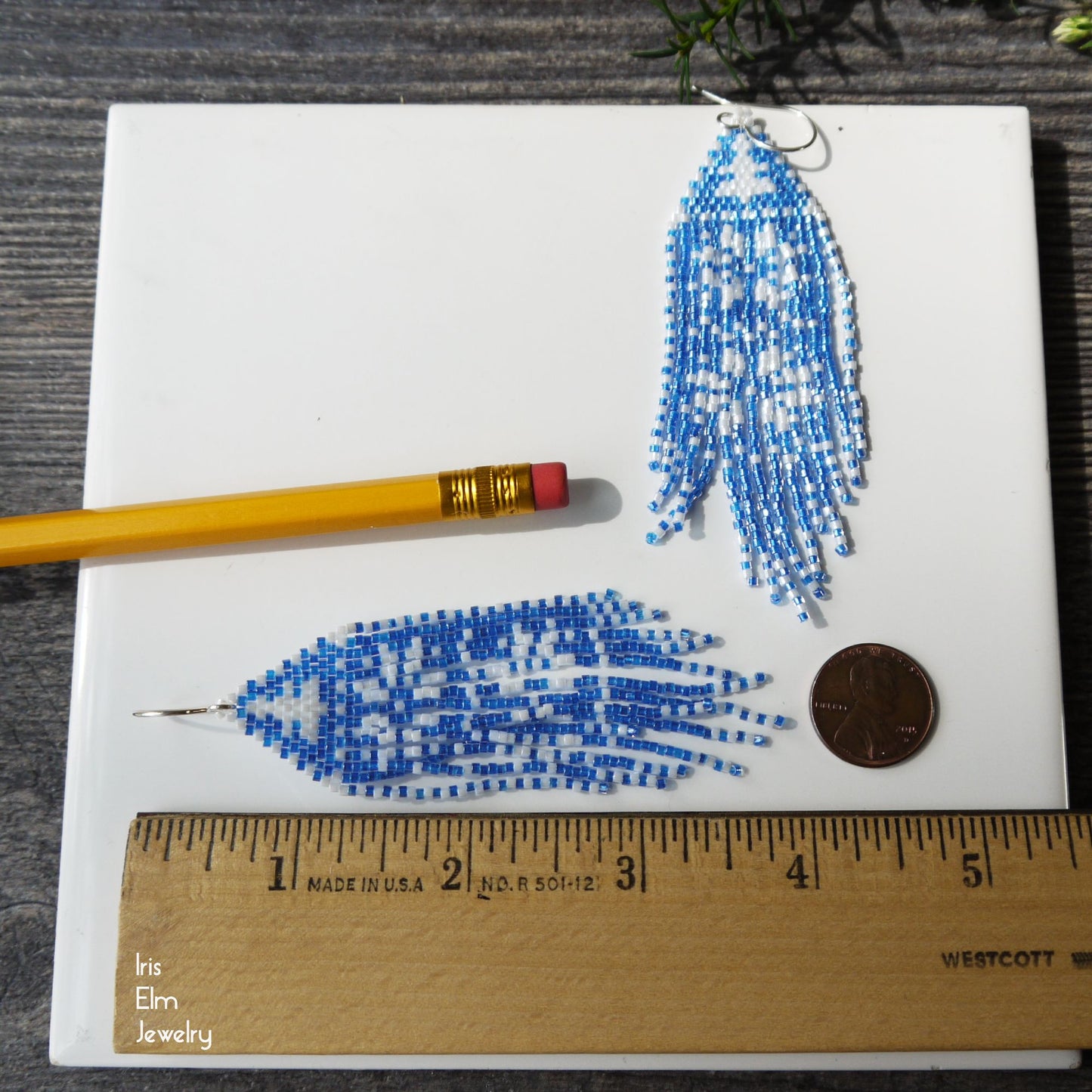4 inch long Blue beaded fringe earrings on a white card with a pencil and coin for scale, on a wooden surface.