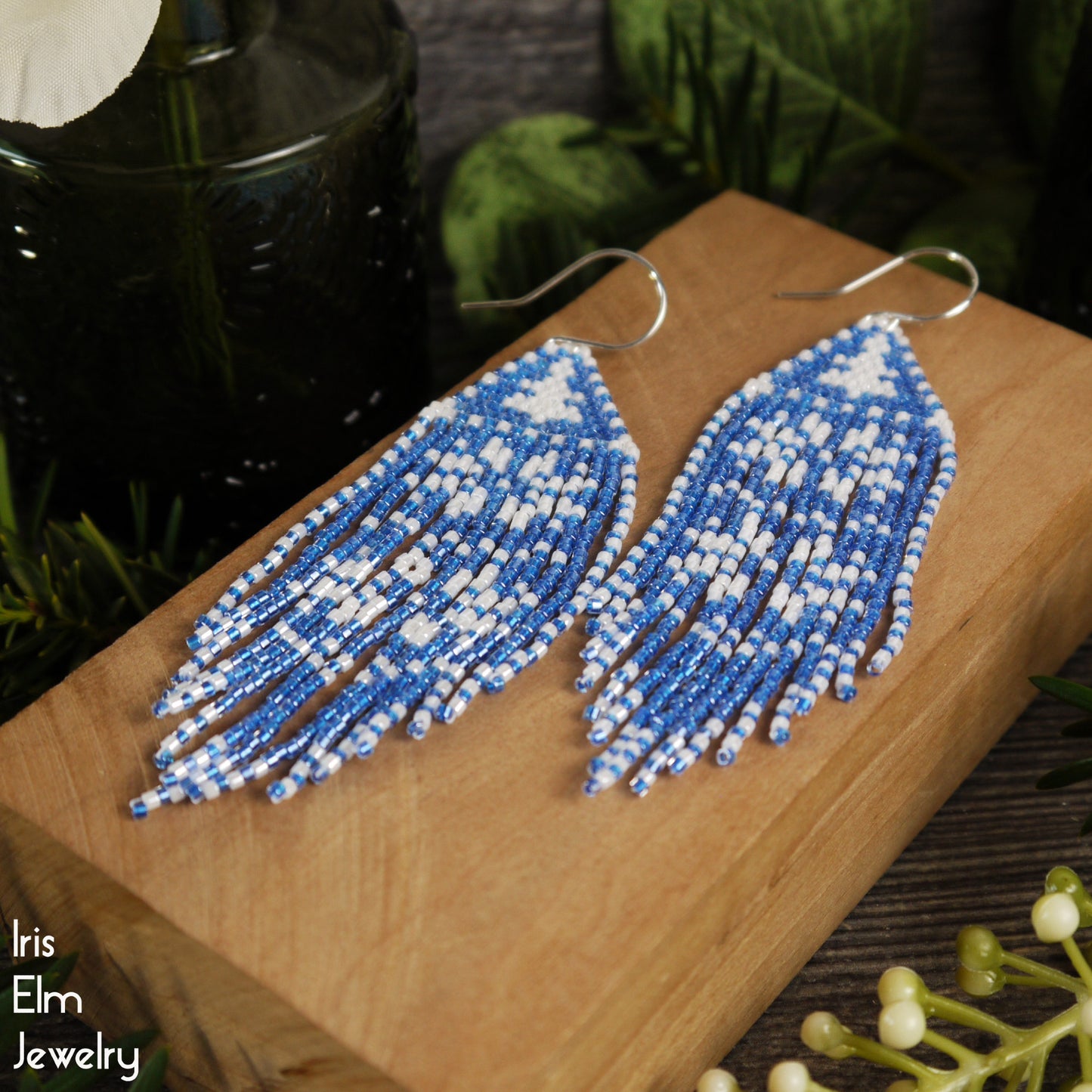 Blue and white beaded snowflake pattern fringe seed bead earrings on a wooden surface with 'Iris Elm Jewelry' branding.