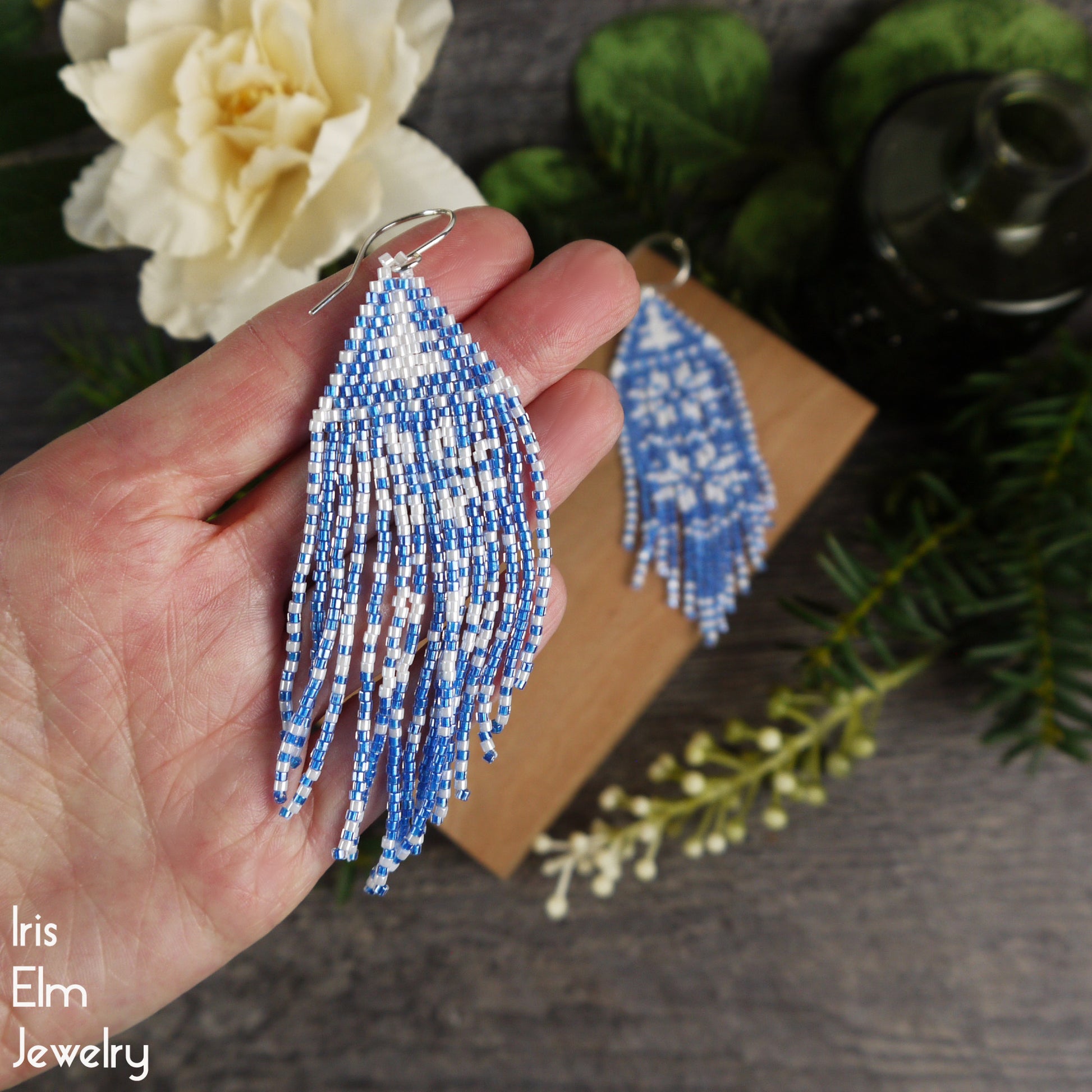 Blue and white beaded snowflake fringe earrings held in a hand with a blurred background