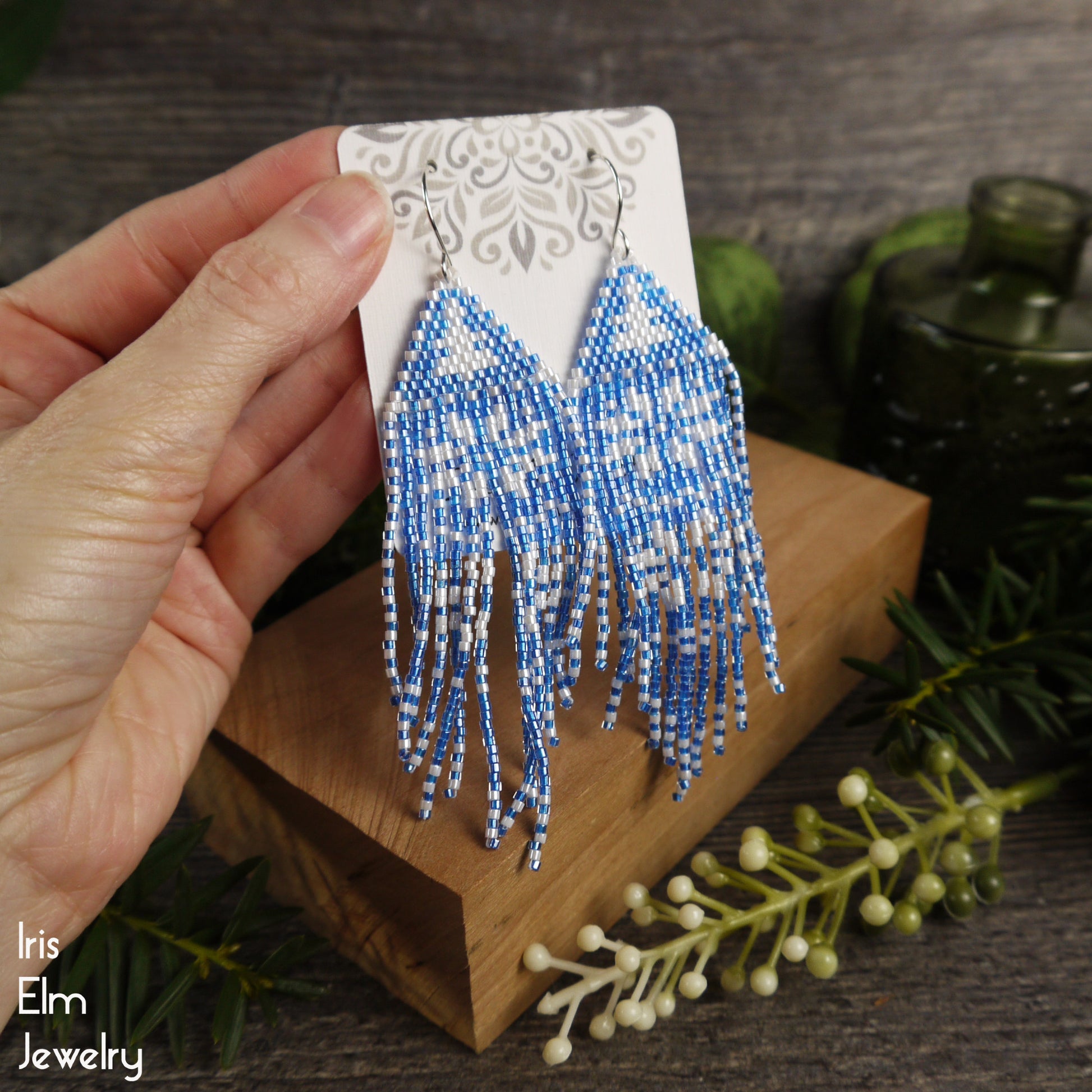 Blue beaded fringe earrings on a wooden block with a blurred background