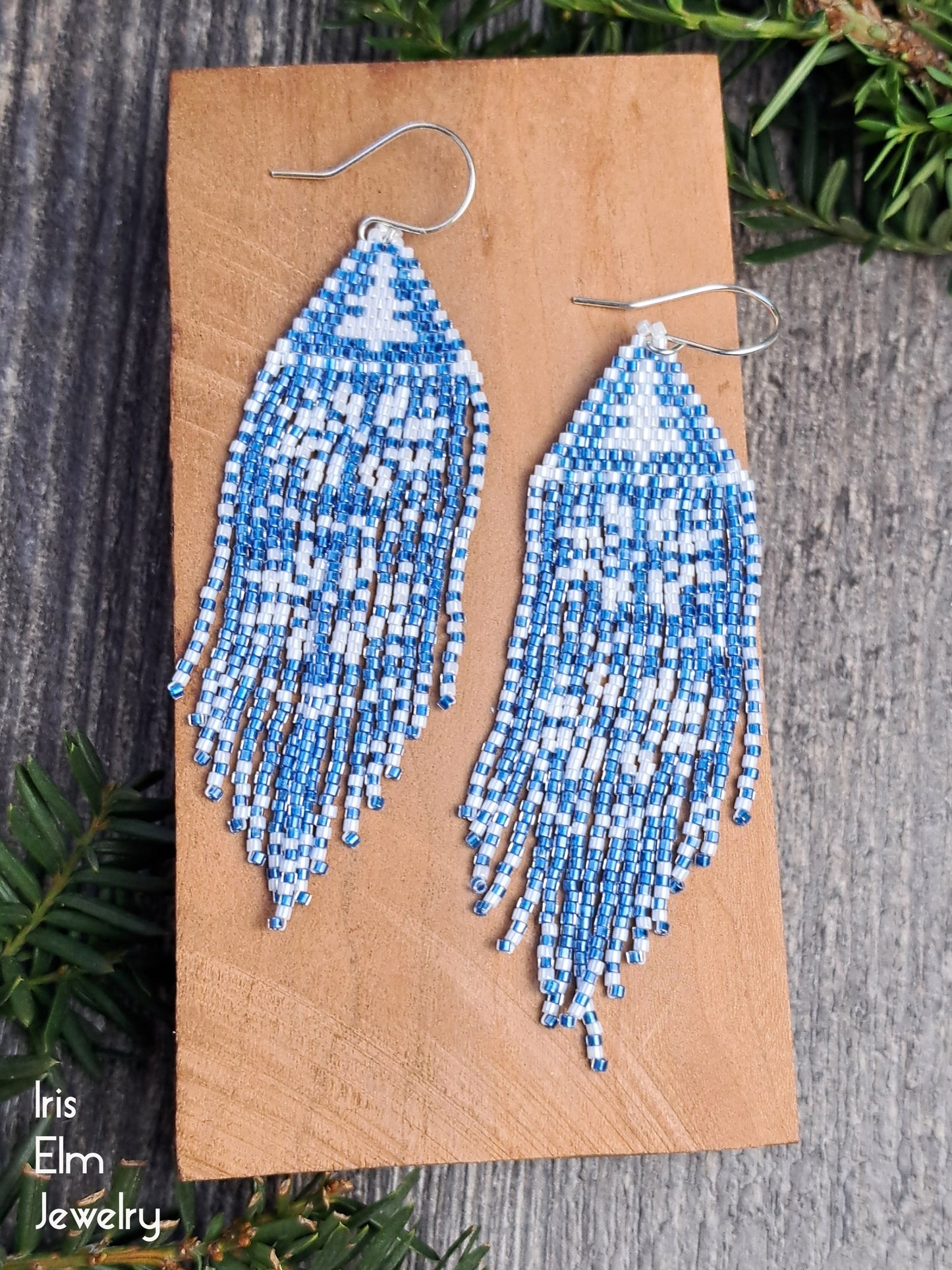 Blue and white beaded winter snowflake fringe seed bead earrings on a wooden card with 'Iris Elm Jewelry' branding.