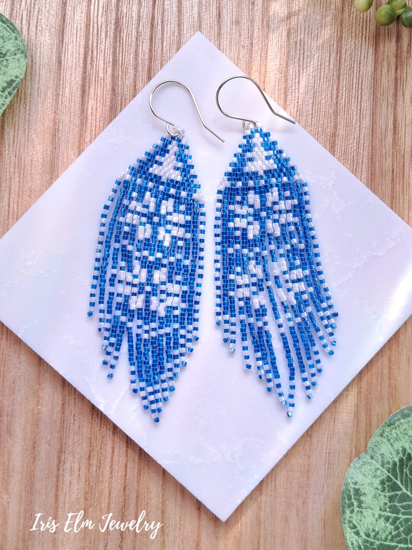 Blue and white beaded snowflake seed bead fringe earrings on a white card with 'Iris Elm Jewelry' branding, on a wooden surface.