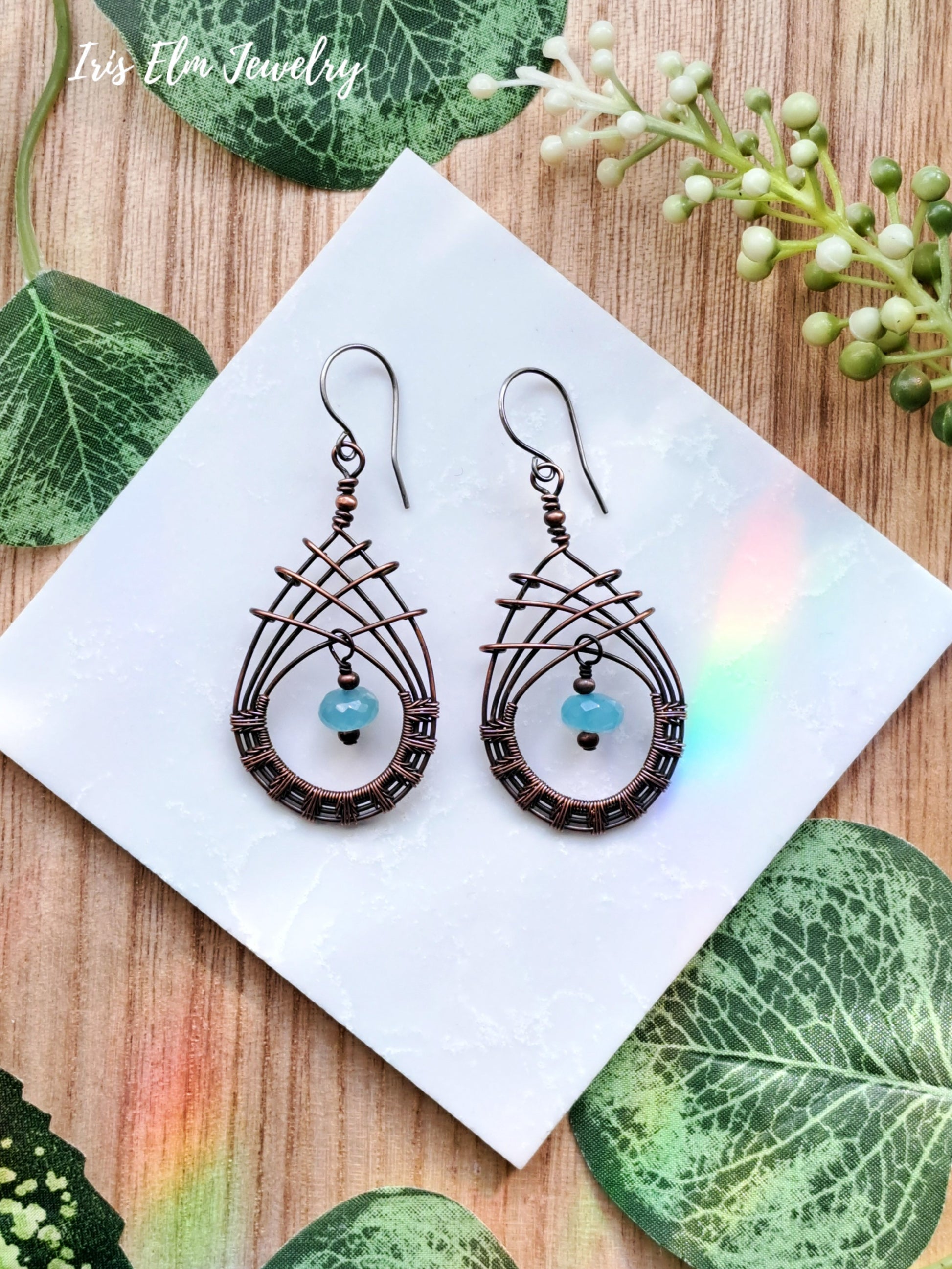 Pair of copper wire woven teardrop blue chalcedony earrings on a white square with green leaves and flowers in the background