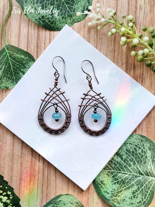 Pair of copper wire woven teardrop blue chalcedony earrings on a white square with green leaves and flowers in the background