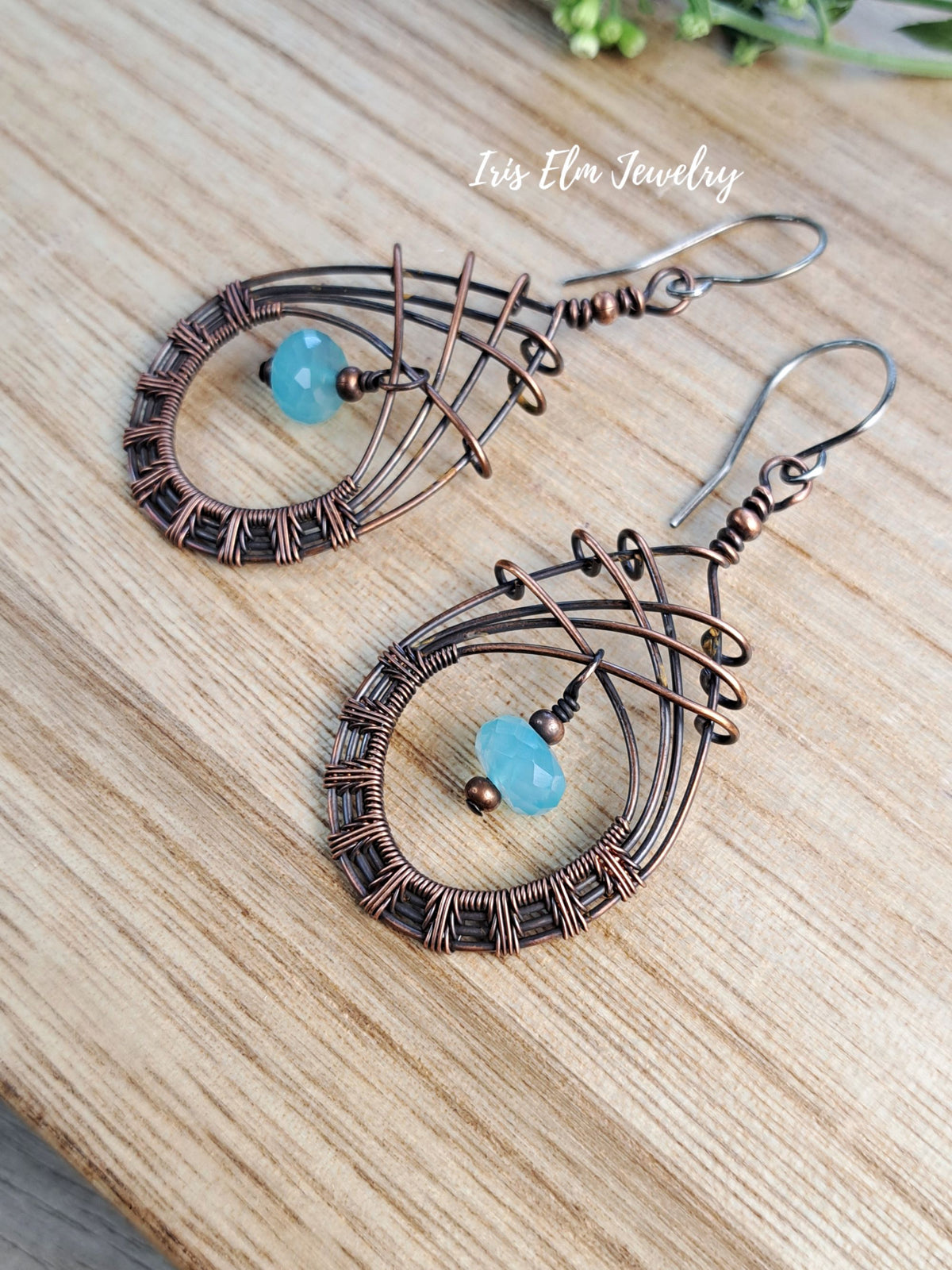 A pair of handmade teardrop-shaped wire-wrapped earrings with a small light blue bead, displayed on a light wood surface. The brand name 'Iris Elm Jewelry' is watermarked near the top.