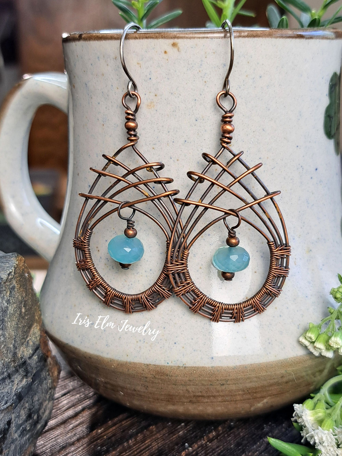 A pair of handmade copper wire dangle earrings with light blue gemstone drops, hanging from a speckled beige ceramic mug on a rustic wooden surface.