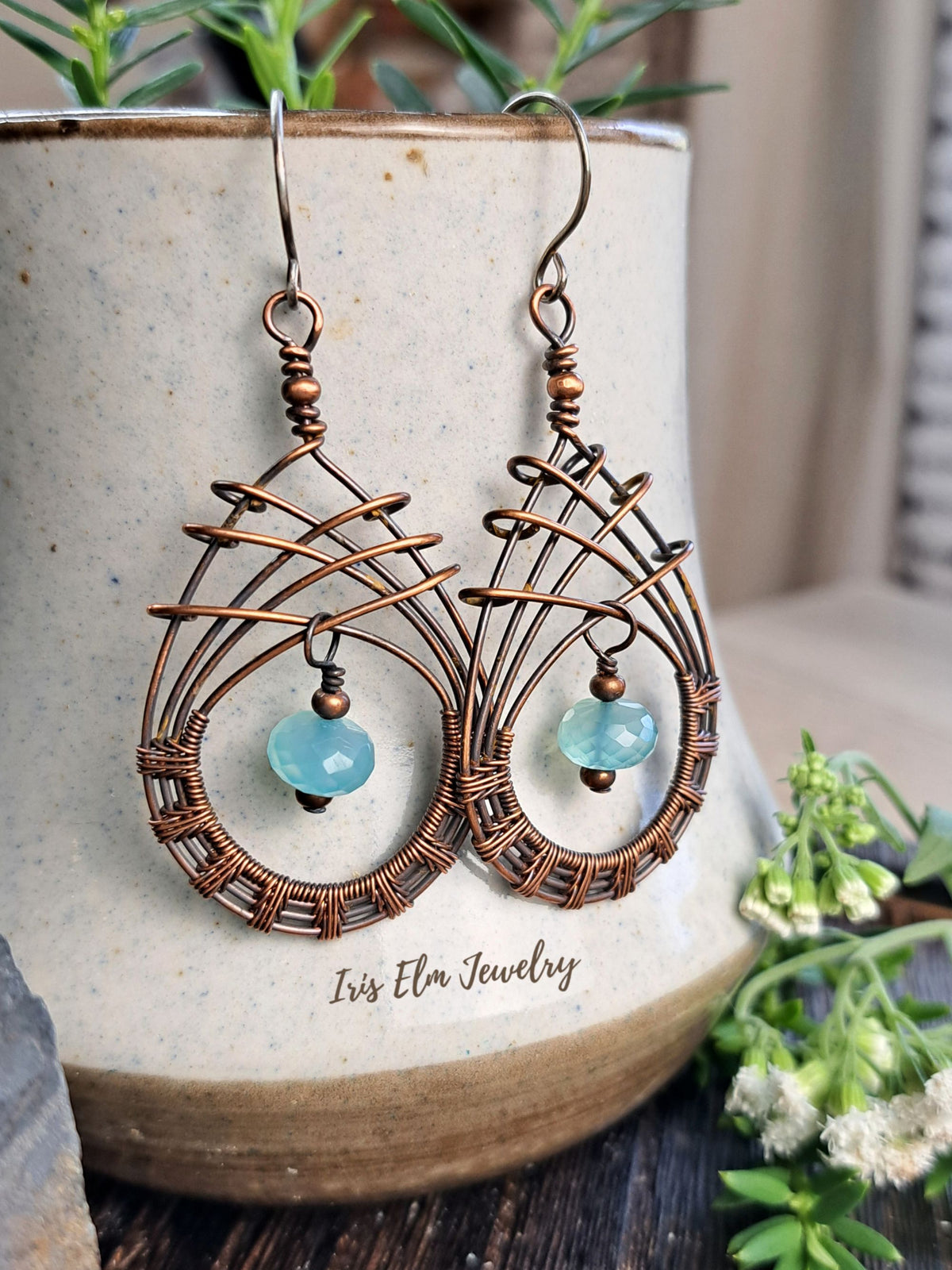 A pair of handmade copper wire dangle earrings with light blue bead chalcedony gemstones, hanging from a ceramic pot with green foliage in the background. The brand name 'Iris Elm Jewelry' is printed on the pot.