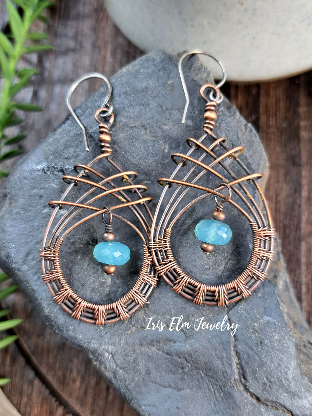 A pair of handmade copper wire dangle earrings with small light blue faceted beads, displayed on a dark gray stone with a wooden surface and greenery in the background. The brand name 'Iris Elm Jewelry' is watermarked at the bottom.