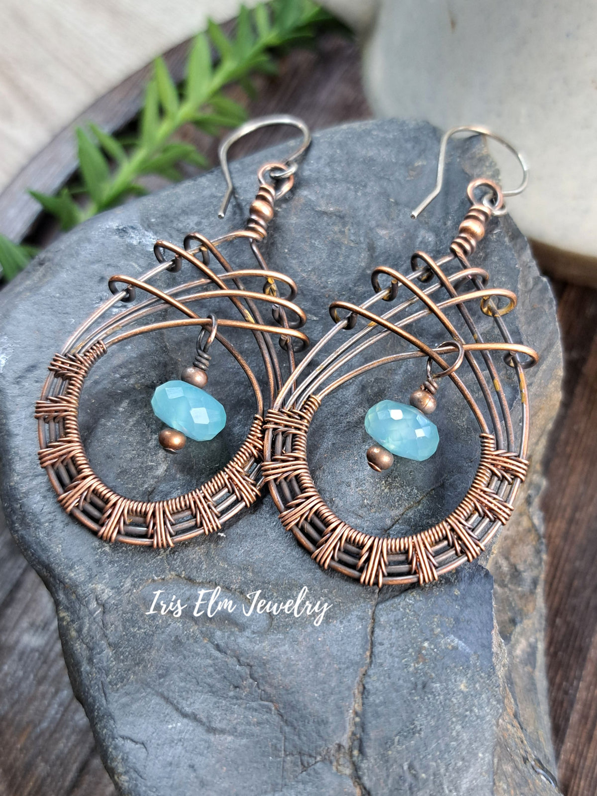 A pair of handmade copper wire hoop earrings with a small faceted light blue bead dangling from each hoop, displayed on a dark gray stone. The brand name 'Iris Elm Jewelry' is watermarked near the bottom.