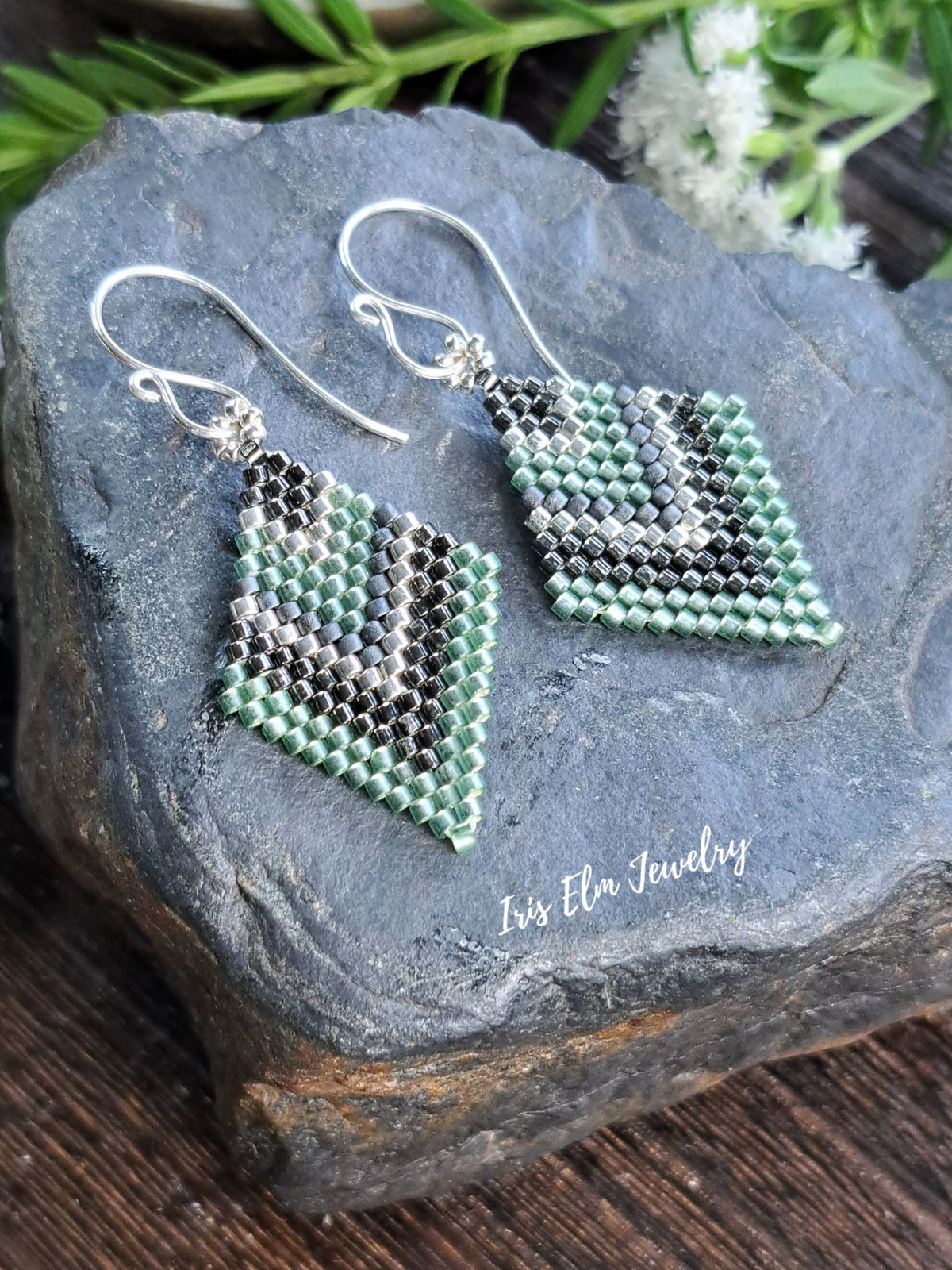 A pair of handmade beaded dangle earrings with a geometric pattern in mint green, black, and white, suspended from silver fishhook wires. The earrings are displayed on a dark gray slate stone with a wooden surface and green foliage in the background.