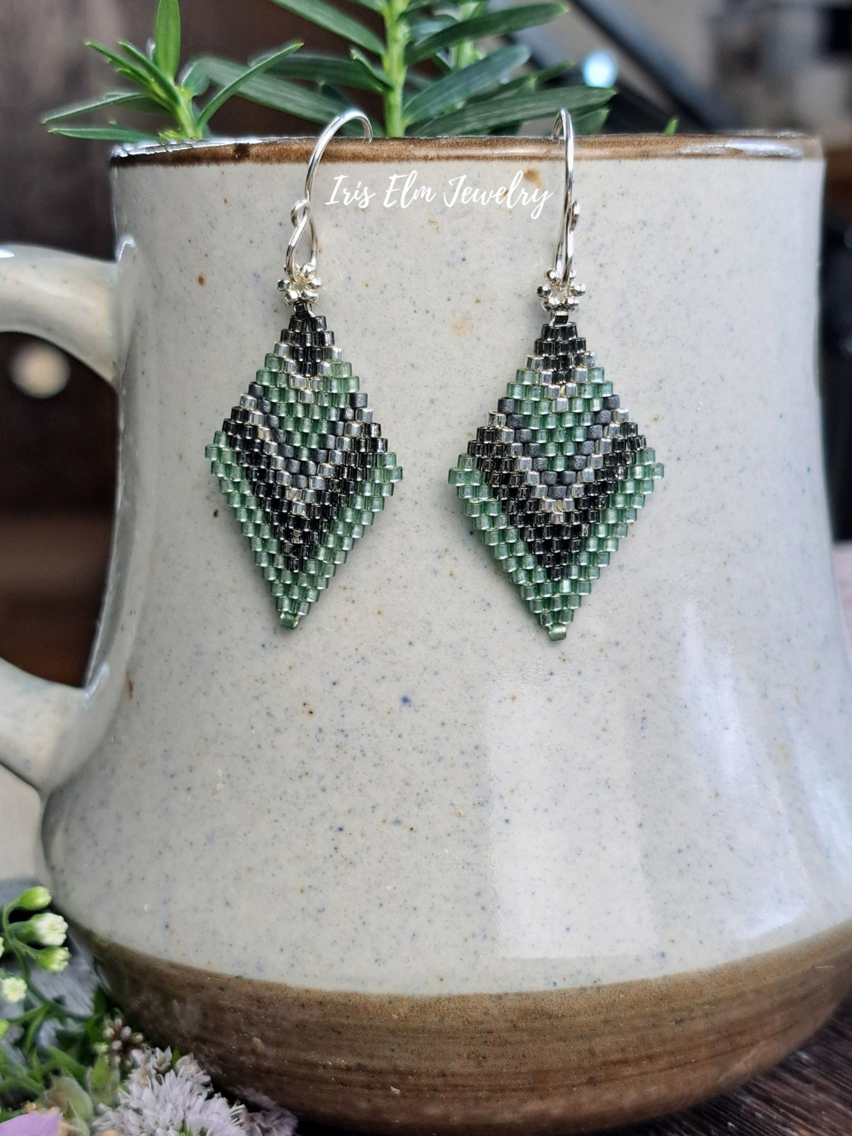 A pair of handmade beaded dangle earrings with a green, grey, and white geometric pattern, hanging from silver hooks in front of a speckled ceramic mug with a rosemary sprig on top.