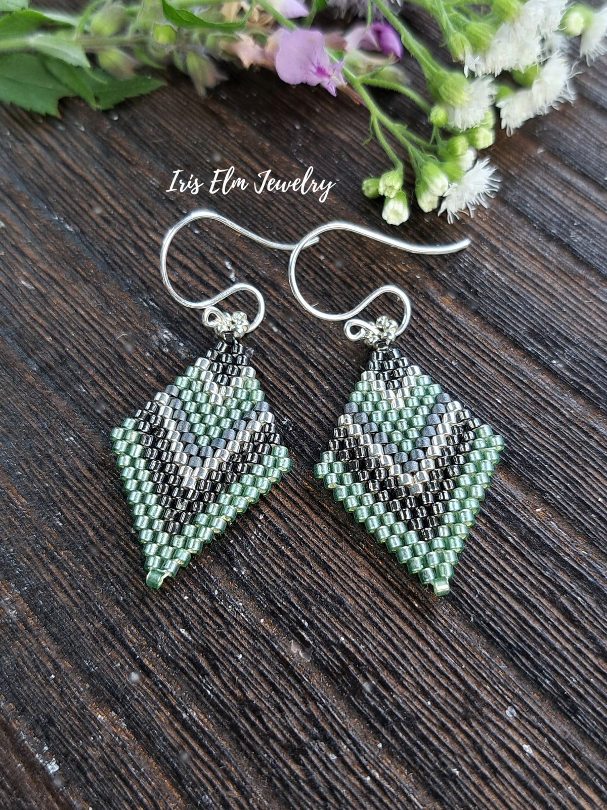 A pair of handmade beaded dangle earrings with a chevron pattern in sage green, grey, and silver, suspended from silver fishhook wires. The earrings are displayed on a dark wood surface with small wildflowers in the background and the text 'Iris Elm Jewelry' written above.