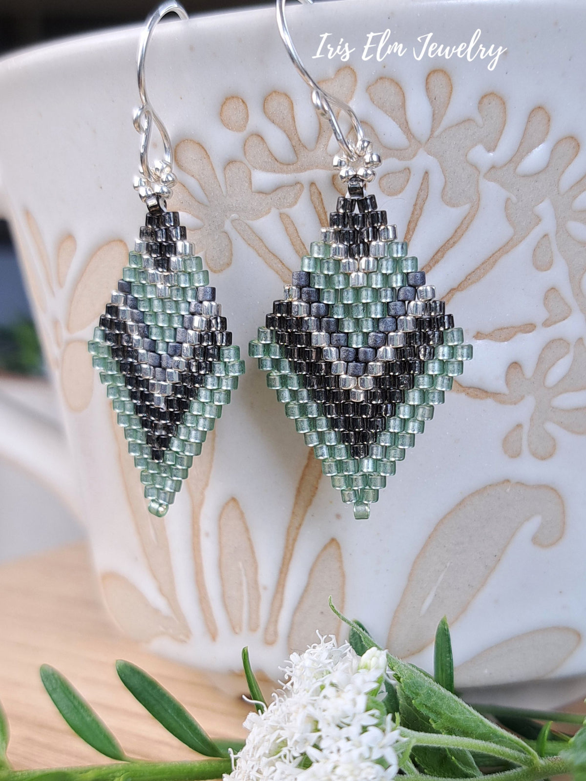 A pair of beaded dangle earrings with a geometric diamond design in sage green, grey, and silver, hanging in front of a white ceramic mug with a floral pattern. The text 'Iris Elm Jewelry' is visible in the top right corner.