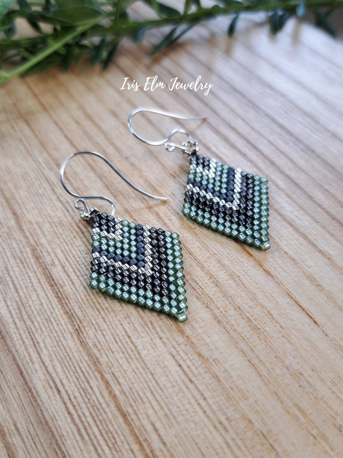 A pair of handmade beaded dangle earrings with a green, grey, and silver seed bead geometric pattern, suspended from silver fishhook wires. The earrings are displayed on a light wood grain surface with a small sprig of green foliage in the background.