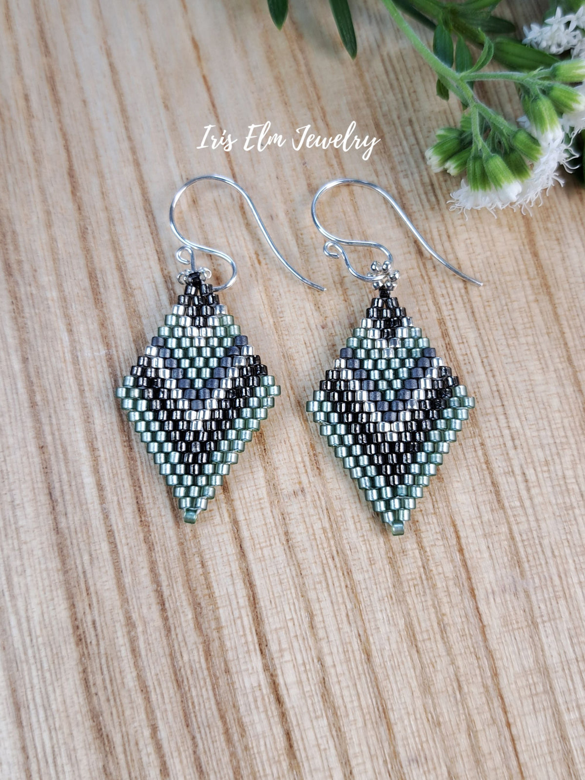 A pair of handmade beaded dangle earrings with a geometric diamond pattern in silver, gray, and mint green, suspended from silver fishhook wires. The earrings are displayed on a light wood grain surface with a small white flower and green leaves in the top right corner.